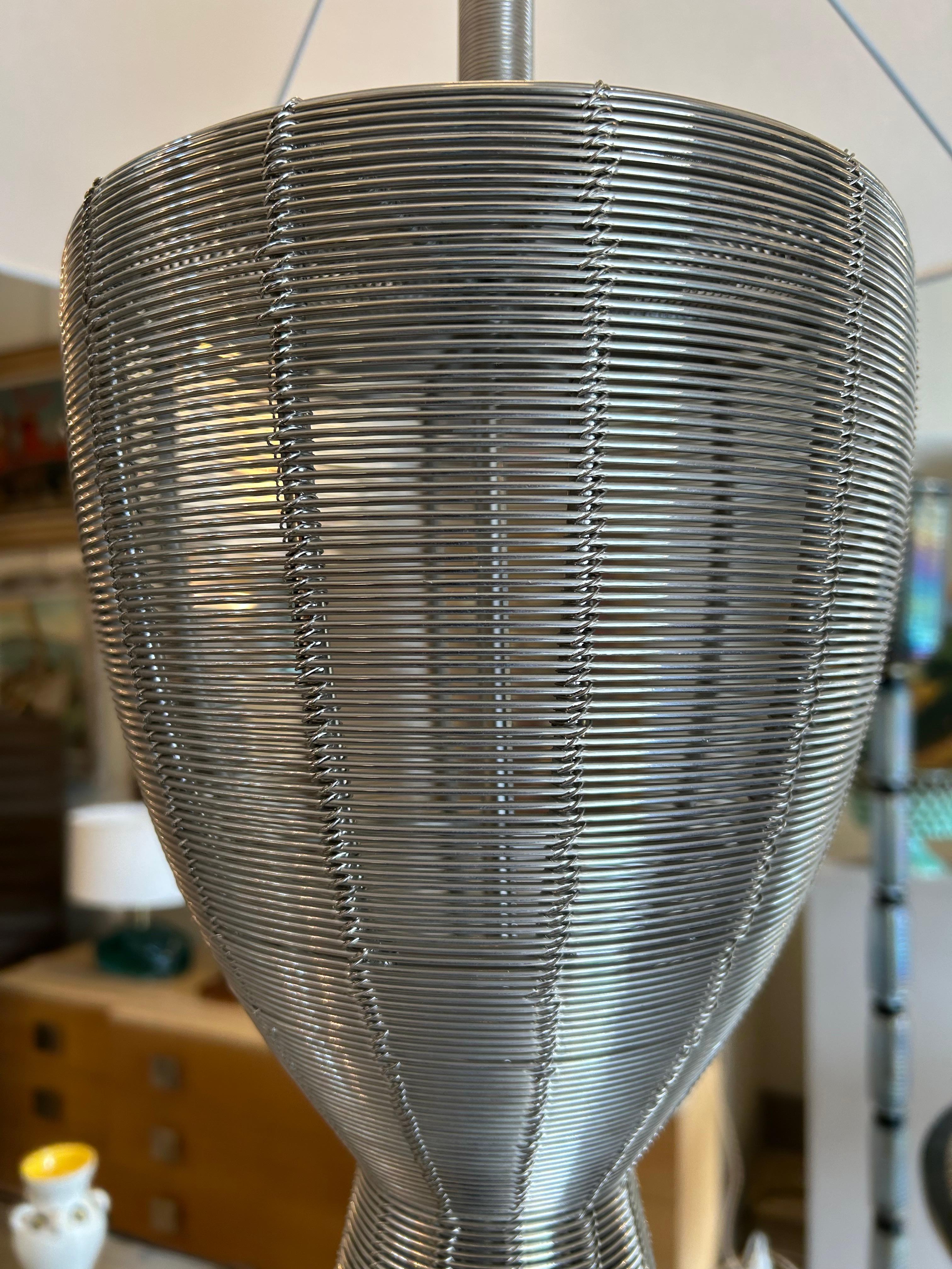 This lamp is a piece of Design in braided metal wire, probably stainless steel. The technique used is that of weaving iron wrapped around a structure, which gives it an airy and sculptural appearance.
It could be inspired by the work of contemporary