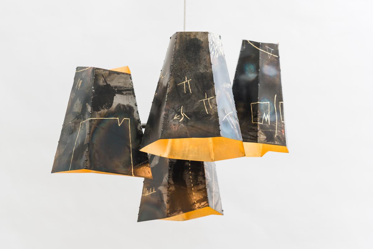 Four hand-built steel shades suspend in the air at various heights creating an organic cluster of asymmetric forms. Each shade is cut and welded individually, before patinas are applied. An abstract language of semi-pictographic images is incised