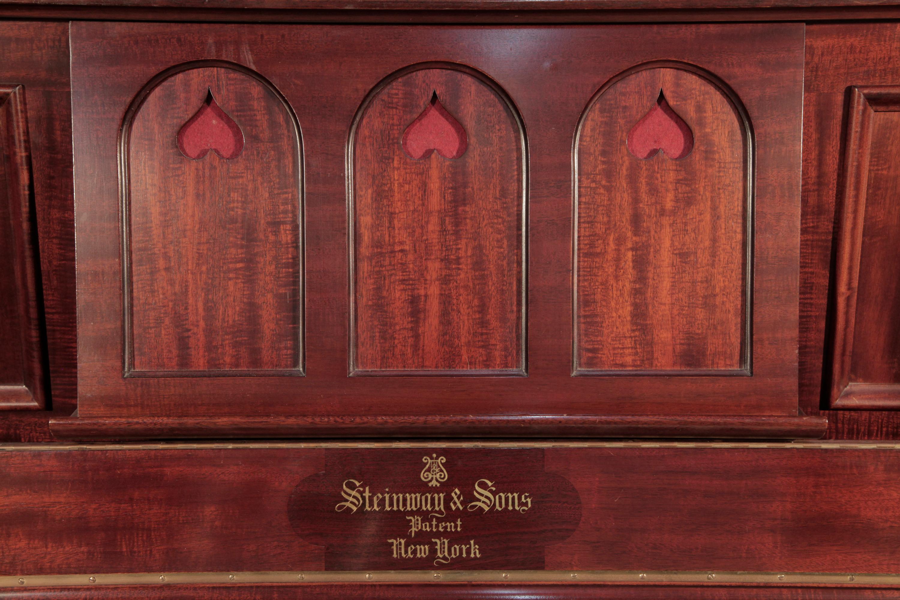Arts and Crafts style, 1905, Steinway vertegrand upright piano with a figured, mahogany case. Cabinet features large sculptural legs with an arched architrave visible from the piano profile. Cabinet features a wide music desk in a three arch design