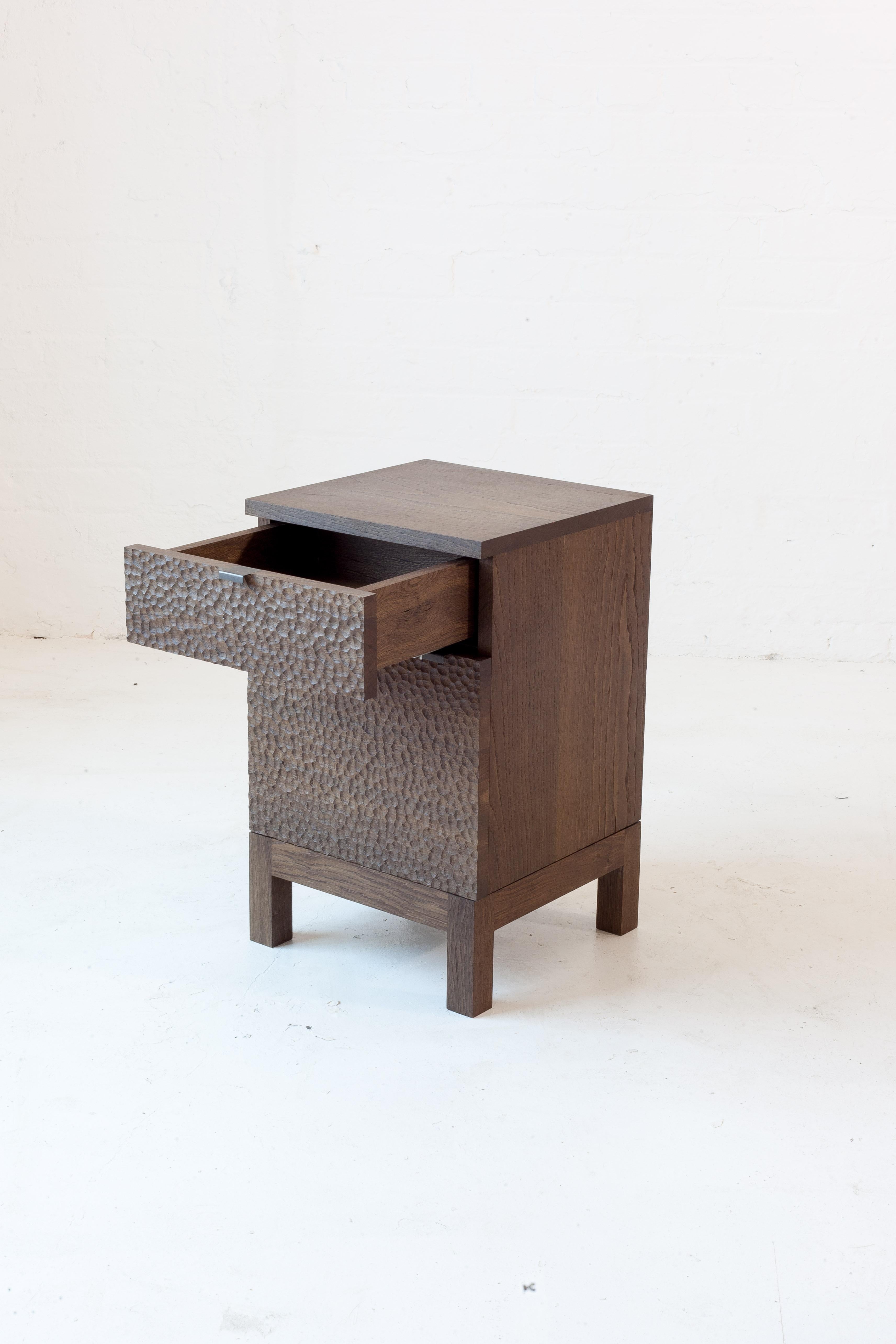 A bedside table with hand-carved textured doors. 

This piece is made in solid English oak. The oak is darkened using the traditional process of fuming, where the timber is exposed to ammonia to react with naturally occurring tannins. This results
