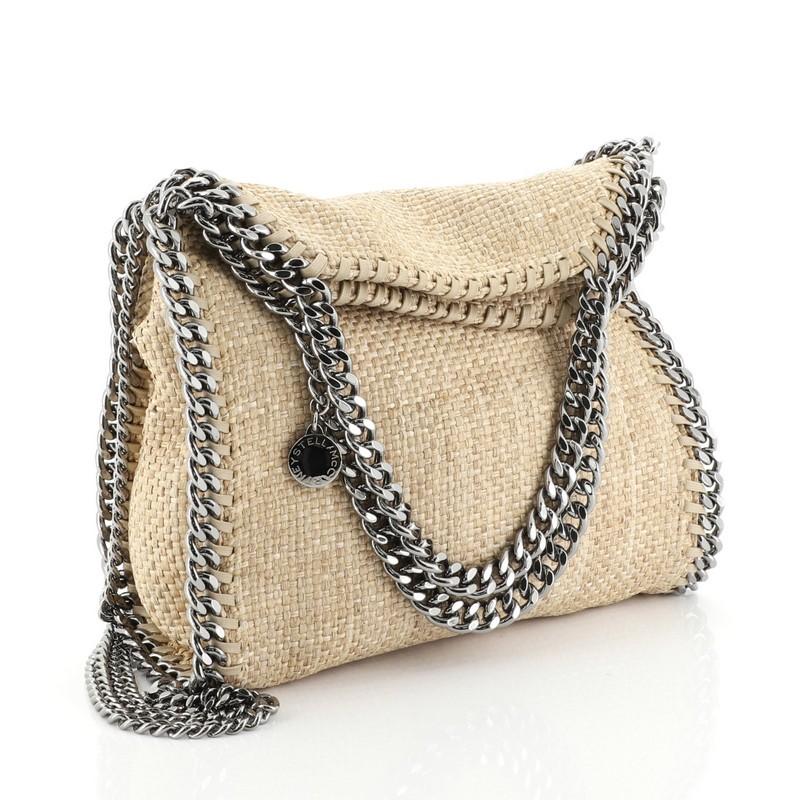 This Stella McCartney Falabella Fold Over Crossbody Bag Raffia Mini, crafted from neutral raffia, features chain link handles and trim, whipstitched edges, and gunmetal-tone hardware. Its magnetic snap closure opens to a black fabric interior with