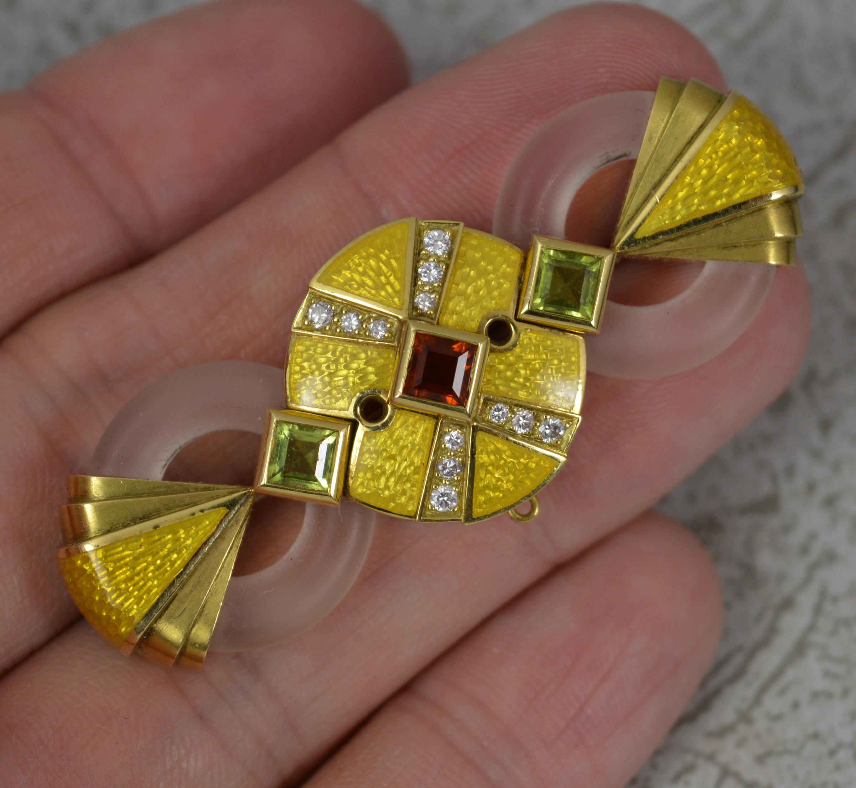 A captivating contemporary brooch of Art Deco design by Stephen Webster.
Modelled in 18 carat yellow gold with a princess cut garnet and peridot to each side. Set with many round brilliant cut diamonds to centre with two circular rock crystal panels