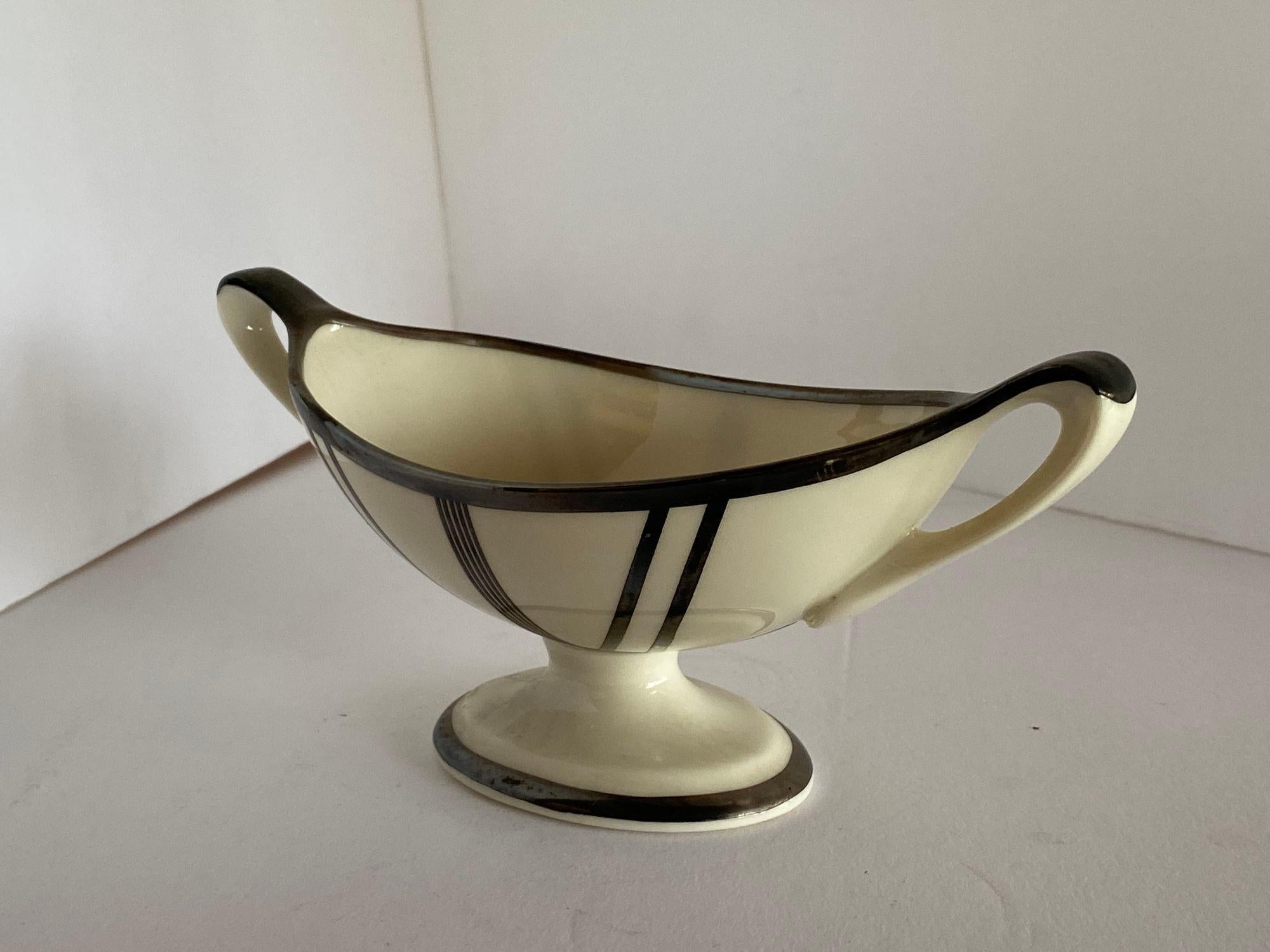 This exquisite sugar bowl, crafted circa 1930, showcases the timeless elegance of Art Deco design with a distinctive stepped silhouette. Manufactured by Lenox, renowned for its fine ceramics, this piece is adorned with a sterling silver overlay,