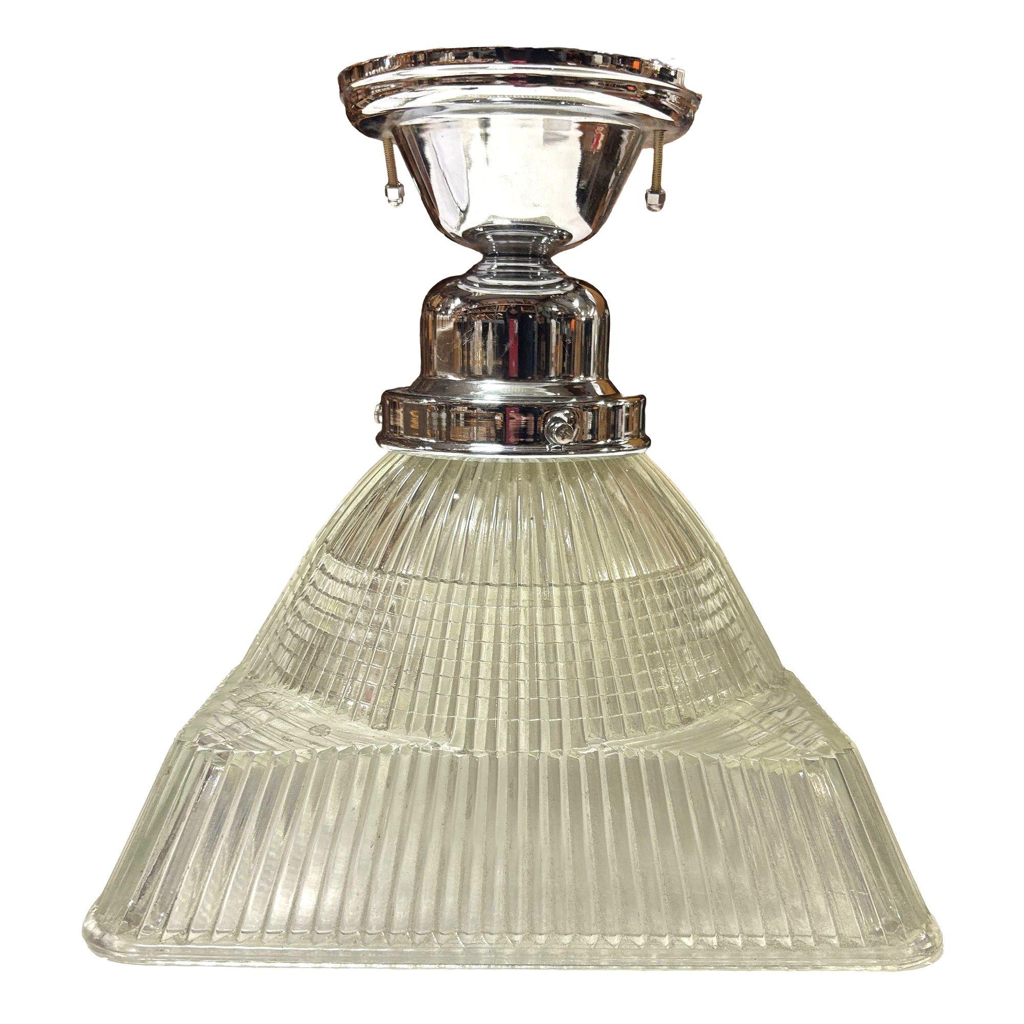 Questa lampada a sospensione presenta un caratteristico paralume quadrato a gradini in vetro Holophane, che unisce estetica industriale e vintage. Il suo vetro prismatico diffonde la luce in modo uniforme, creando una calda luminosità ambientale. È