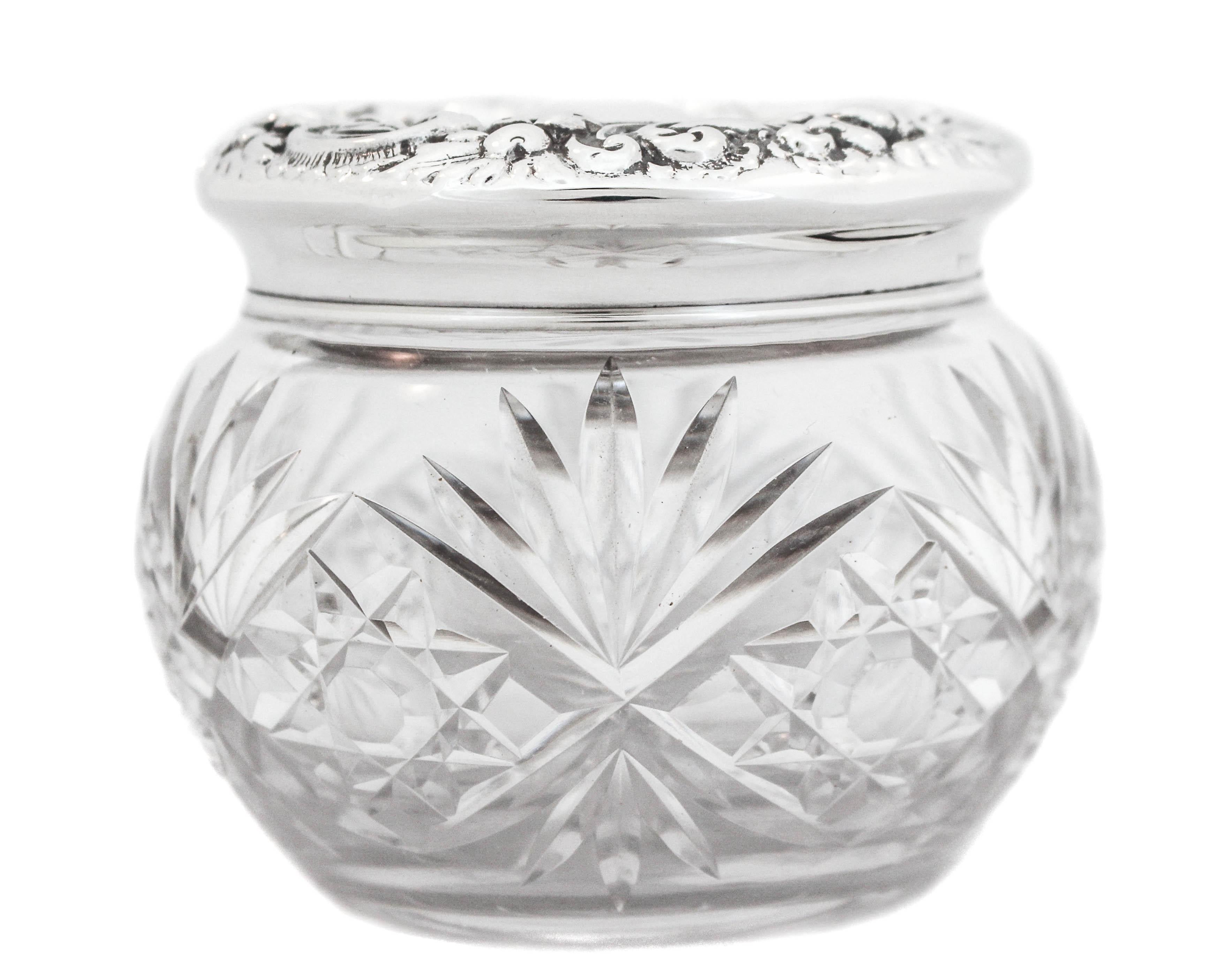 A beautiful sterling silver Repousse lid on a cut-glass jar.  The cover has beautiful blown out flowers and swirls along the edge while in the center there’s a space for a monogram.  The jar has cut glass all around and a starburst on the bottom. 