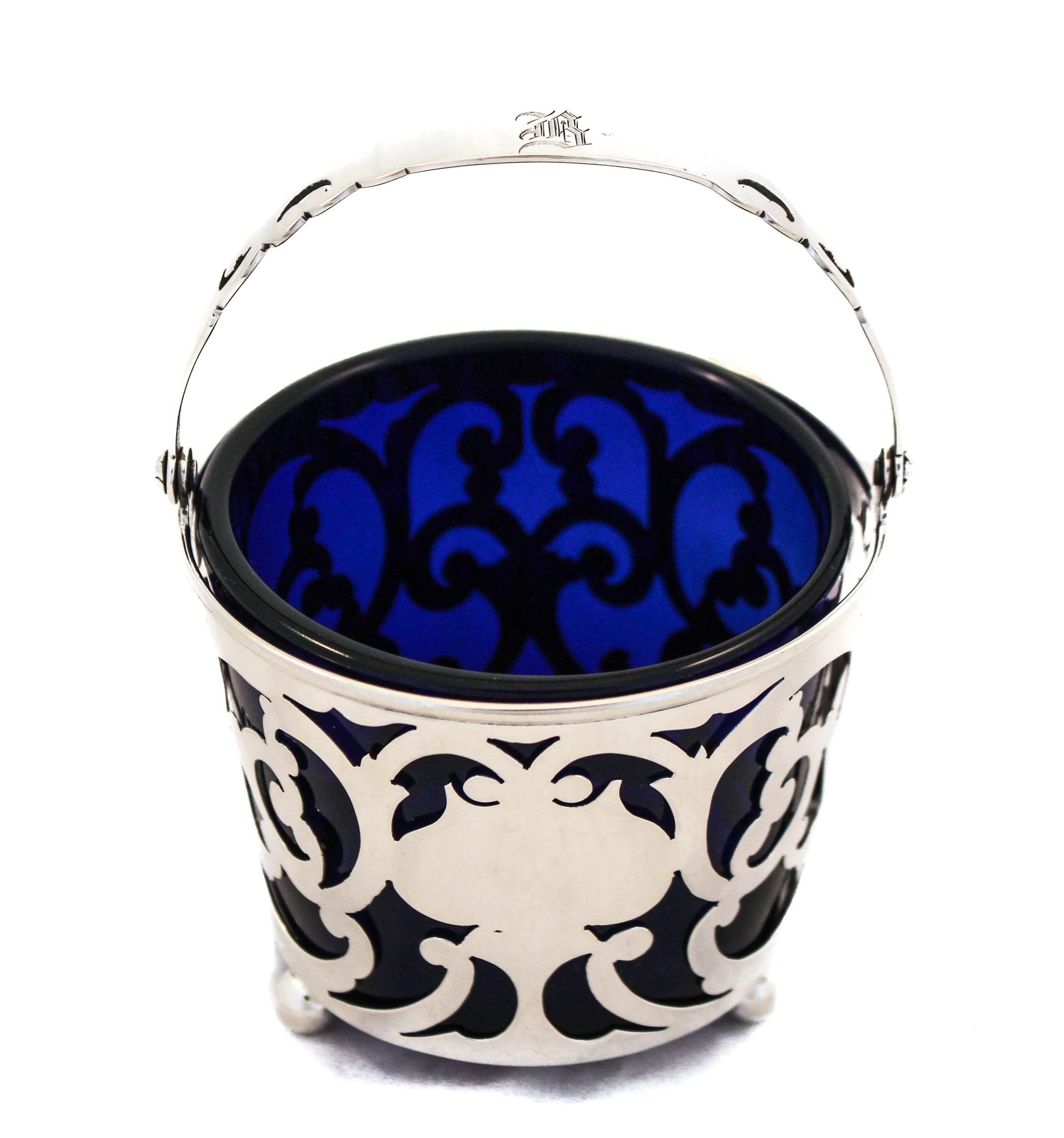 This lovely sterling silver bonbonniere is being offered. Standing on three balls, the entire body is a cutout design. Through the sterling silver the rich blue liner pops out. The handle folds down which makes storing easy. The glass liner comes