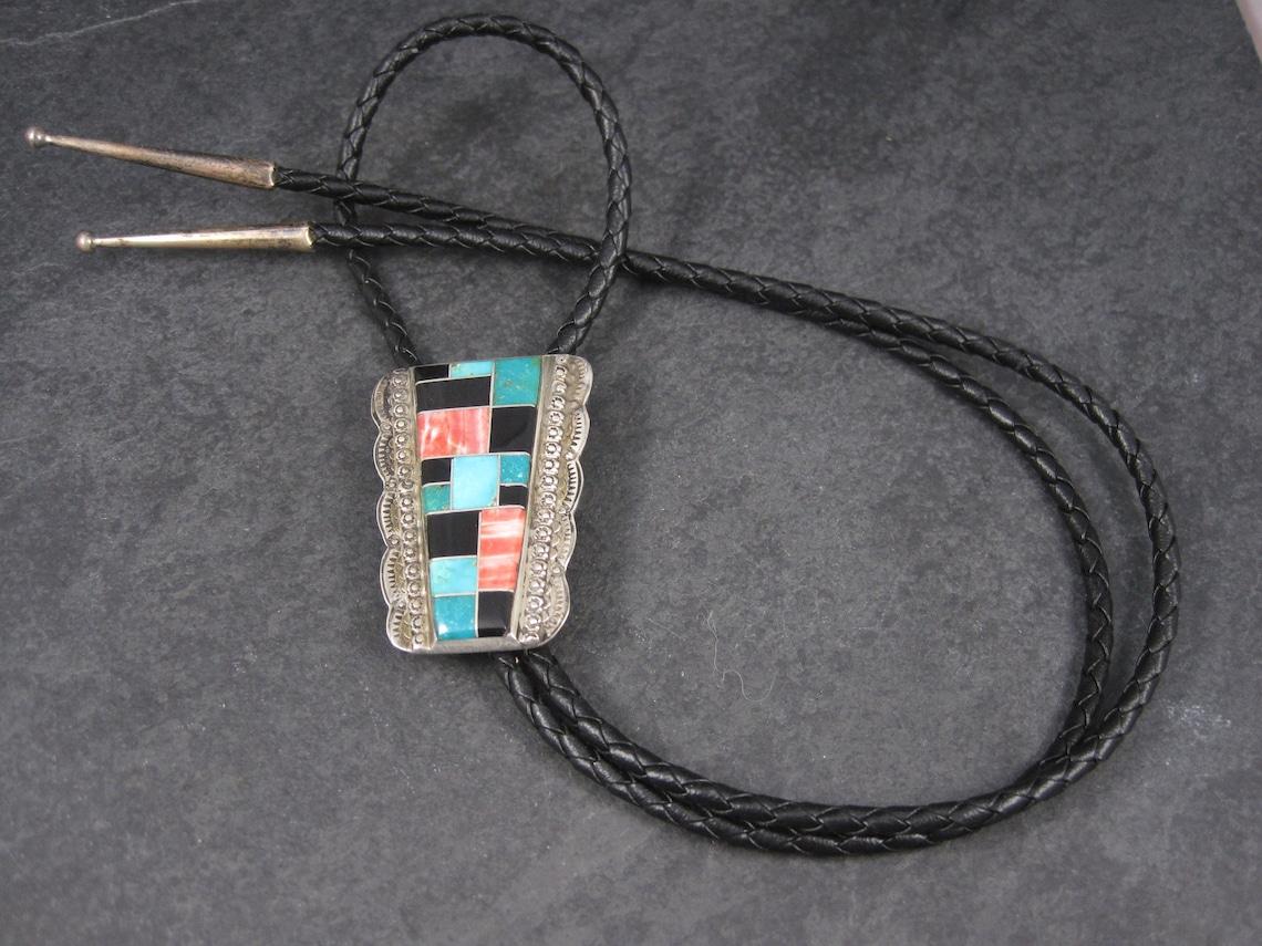 This gorgeous Navajo bolo tie features inlay in blue and green turquoise, spiny oyster and black onyx.

It measures 1 7/16 inches wide and 2 inches long.

Marks: Sterling, F. Tom.

The 37 inch tie is genuine leather, in excellent condition, with 1