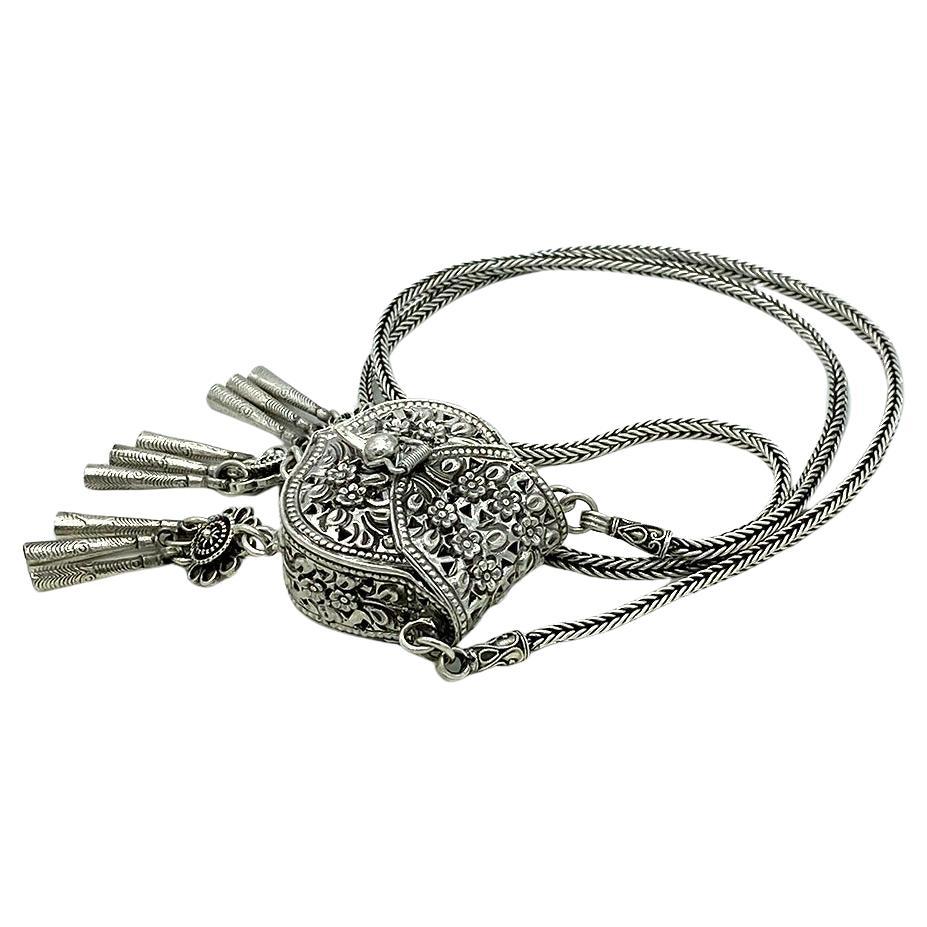 This is a sterling necklace with dangling purse. This 24 inch long necklace is attached to a 1.5 x 3.5 x 1 inch purse pendant. This detailed purse with flower cut out pattern and three groups of dangling fringes also has a hinged lid. Marked 925