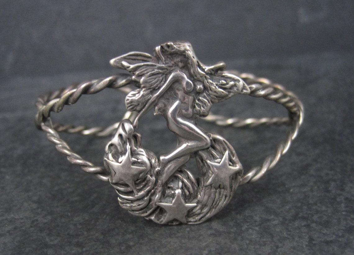 This gorgeous sterling silver cuff bracelet features a nude fairy goddess standing on billowy clouds and stars.

This bracelet measures 1 3/8 inches north to south.
The inner circumference measures 6 1/4 inches including a 5/8 of an inch