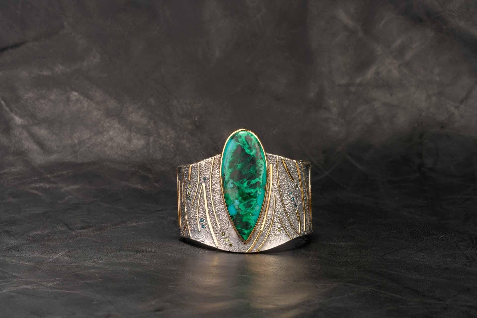 An oxidized sterling silver, 18k yellow gold, and 24k yellow gold featuring one 50x20mm pear shaped cabochon chrysocolla/malachite and 0.535 total carat weight round full cut blue and green diamonds, measures 2.24inches x 1.71inches inside. This