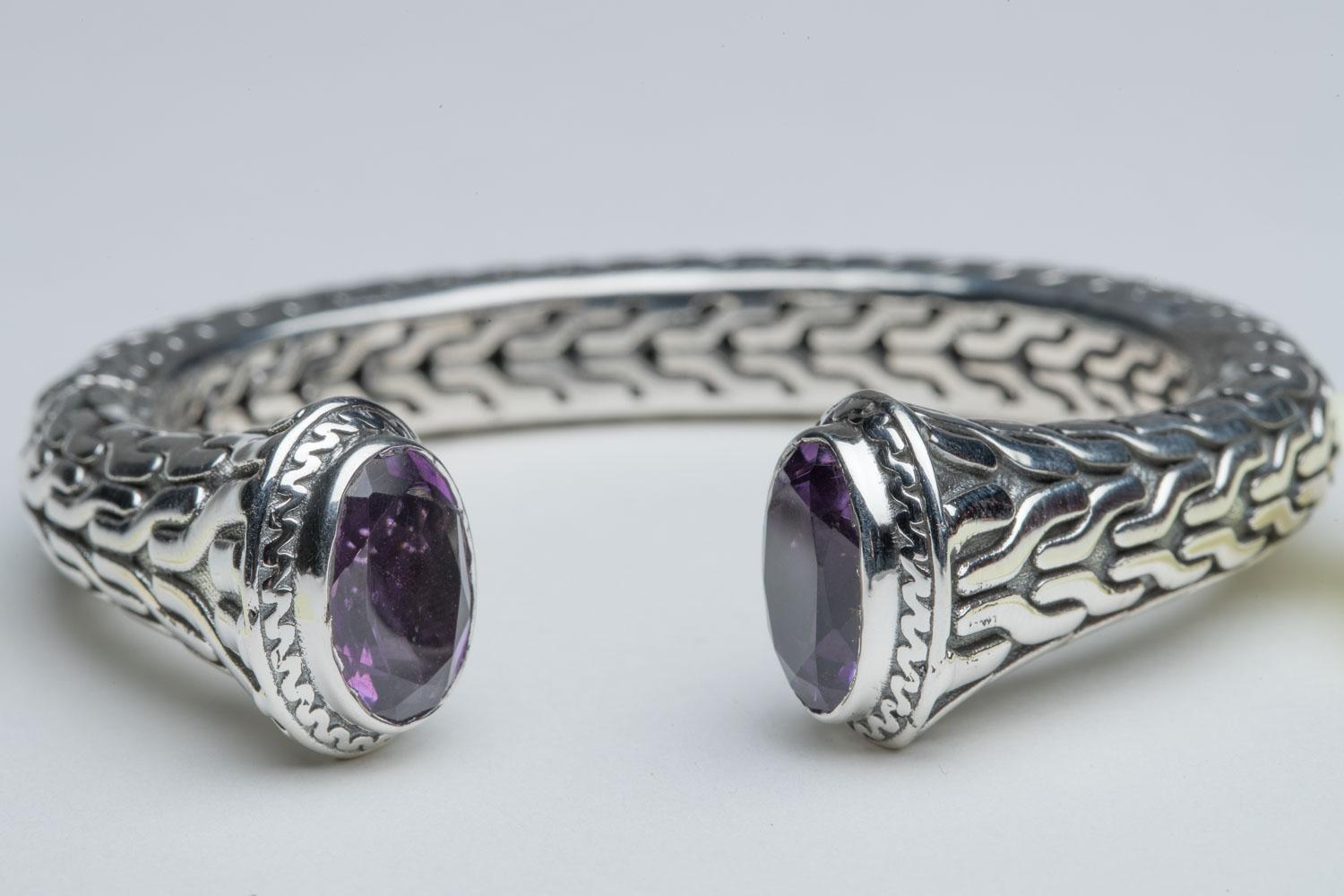 A hand crafted sterling silver clamper or cuff bracelet with a herringbone pattern along the lines of John Hardy.  It is hinged on the side for easy on and off.  The top features two large faceted, oval amethyst stones.  Inside circumference is 6.5