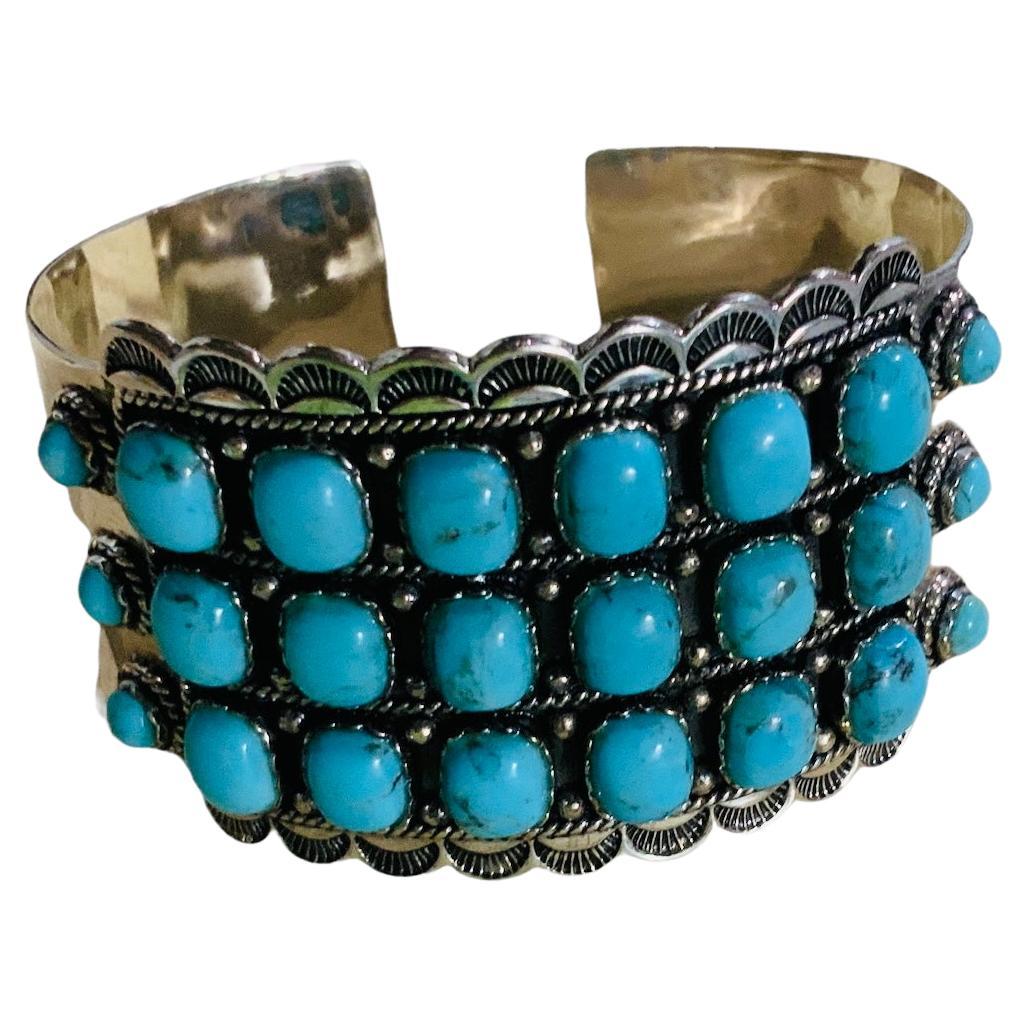 This is a Sterling Silver and Turquoise cuff bracelet. It depicts a wide silver cuff adorned in the front with three rows of six square cabochon turquoises on bezel setting in each one. Twisted ropes and beads enhance each row. At each side of this