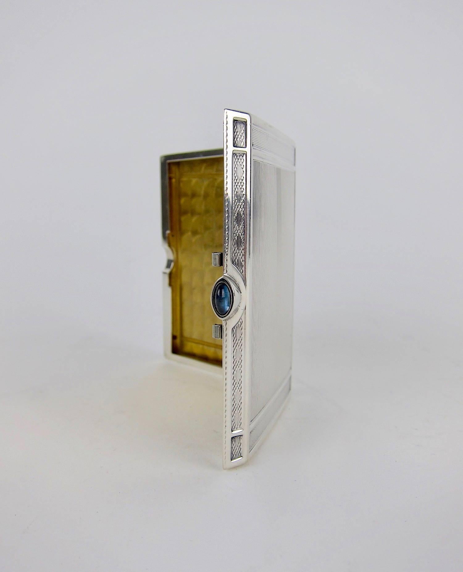 A vintage sterling silver cigarette case with a jeweled cabochon clasp and gilt interior, dating to the late 1940s. The Art Deco silver case was manufactured by Elgin Silversmith Company of New York with elegant, engine-turned decoration; the blue