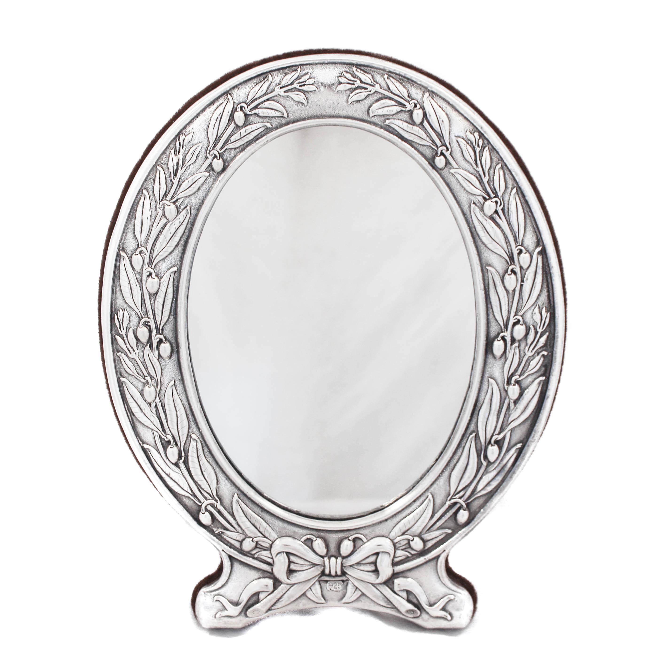 This sterling silver mirror is made in the Art Nouveau style. It is oval and has leaves and flowers going up each side. The silver has a matte finish and is “parentheses” to the mirror. On the bottom there’s a bow tying the two sides together. The