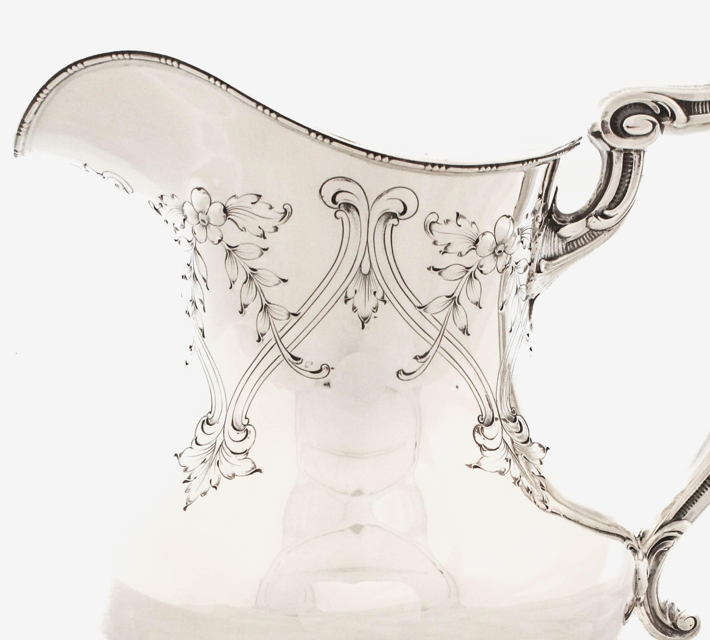 Its sweeping silhouette flows with soft curves from the flared spout down to a wide, ornate base. The handle is especially striking—elaborately cast with swirling, nature-inspired motifs reminiscent of vines or tendrils, adding a sculptural elegance