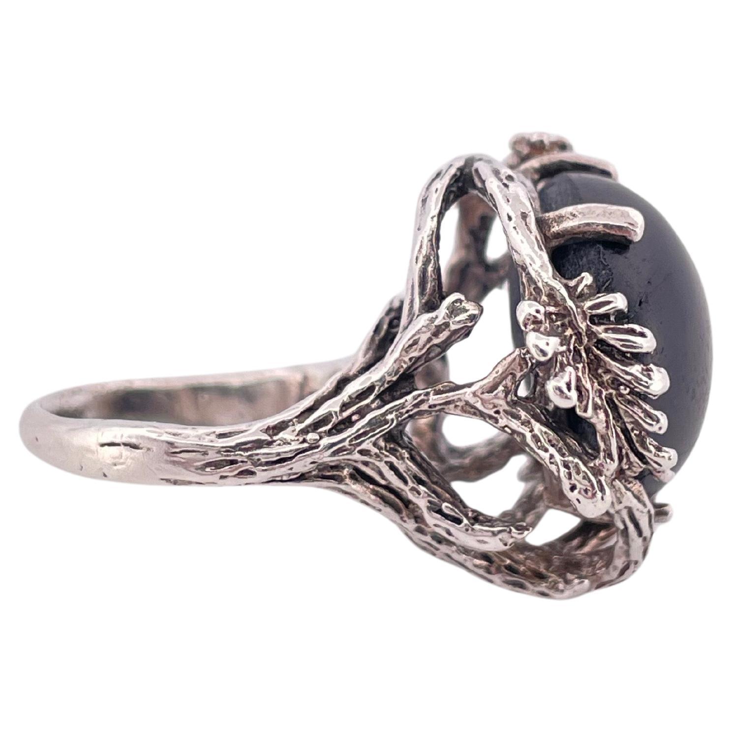 Questo sorprendente anello vintage mette in mostra un affascinante zaffiro nero cabochon, che pesa circa 9,9 grammi in totale. Lo zaffiro presenta un asterismo ipnotico, rivelando un sottile motivo a stella quando viene toccato dalla luce.