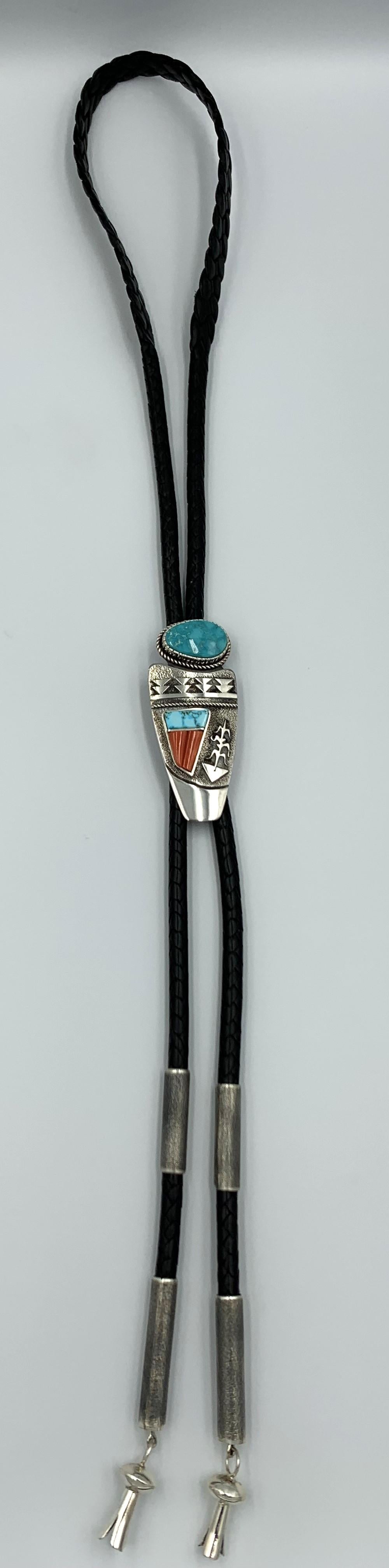 Sterling silver bolo tie by Navajo silversmith Jack Tom. The 1 3/8” x 3 1/4” silver overlay bolo has a 5/8” x 1” Pilot Mountain cabochon along with Blue Gem turquoise and coral overlay. The bolo is on a 46” braided leather strap with silver ends and