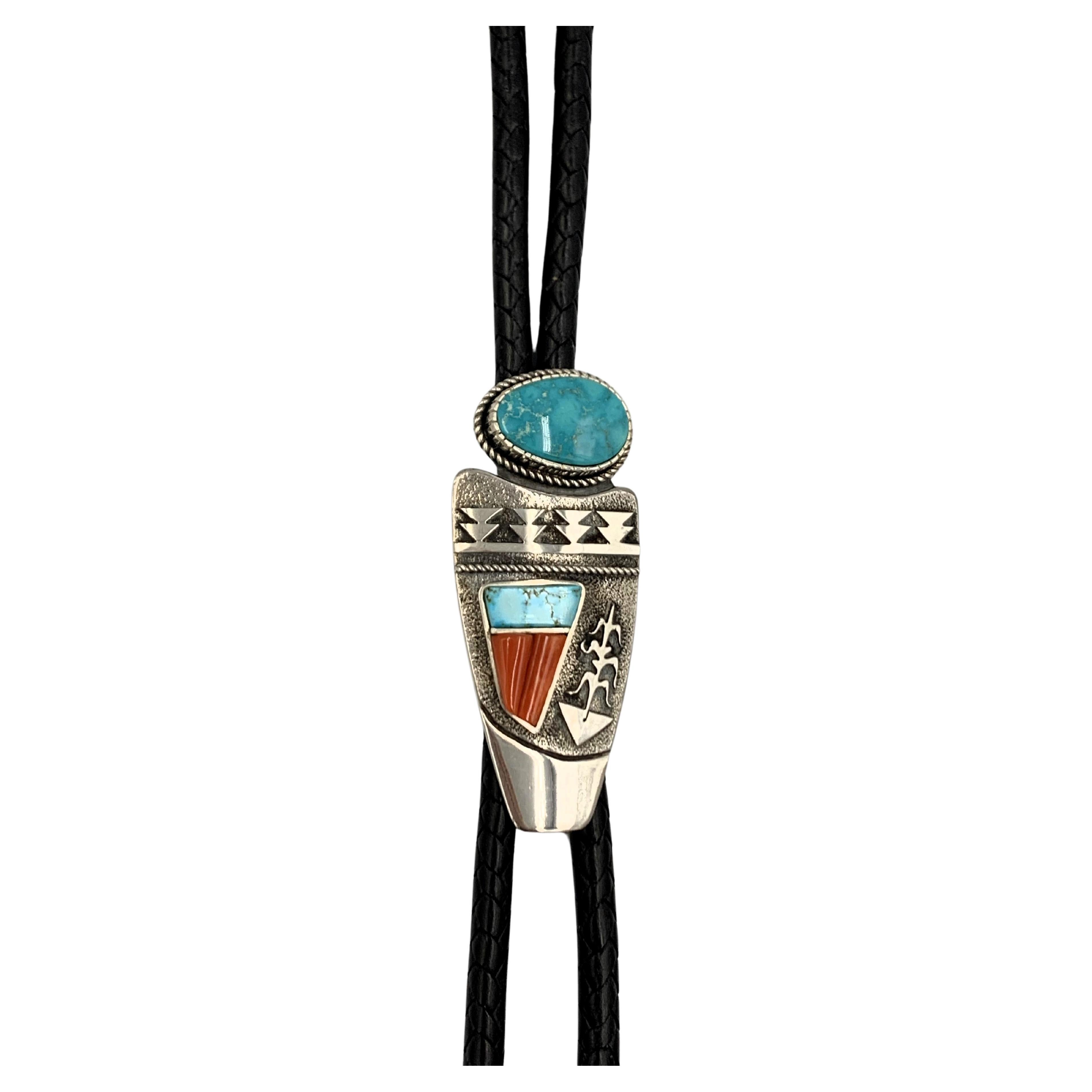 Sterling silver bolo tie by Navajo silversmith Jack Tom. The 1 3/8” x 3 1/4” silver overlay bolo has a 5/8” x 1” Pilot Mountain cabochon along with Blue Gem turquoise and coral overlay. The bolo is on a 46” braided leather strap with silver ends and
