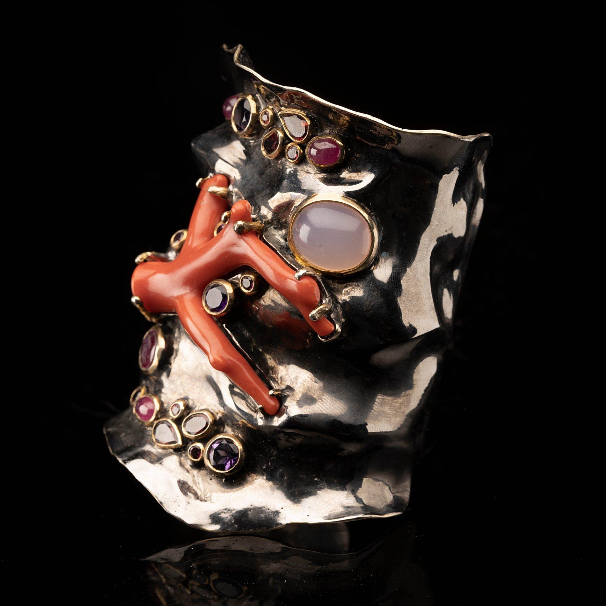 This showstopping number features a red coral branch set centrally in a rhodium plated sterling silver adjustable cuff bracelet set off by a round rose quartz cabochon along with sparkling garnets, sapphires, rubies, amethysts, and 14 karat gold
