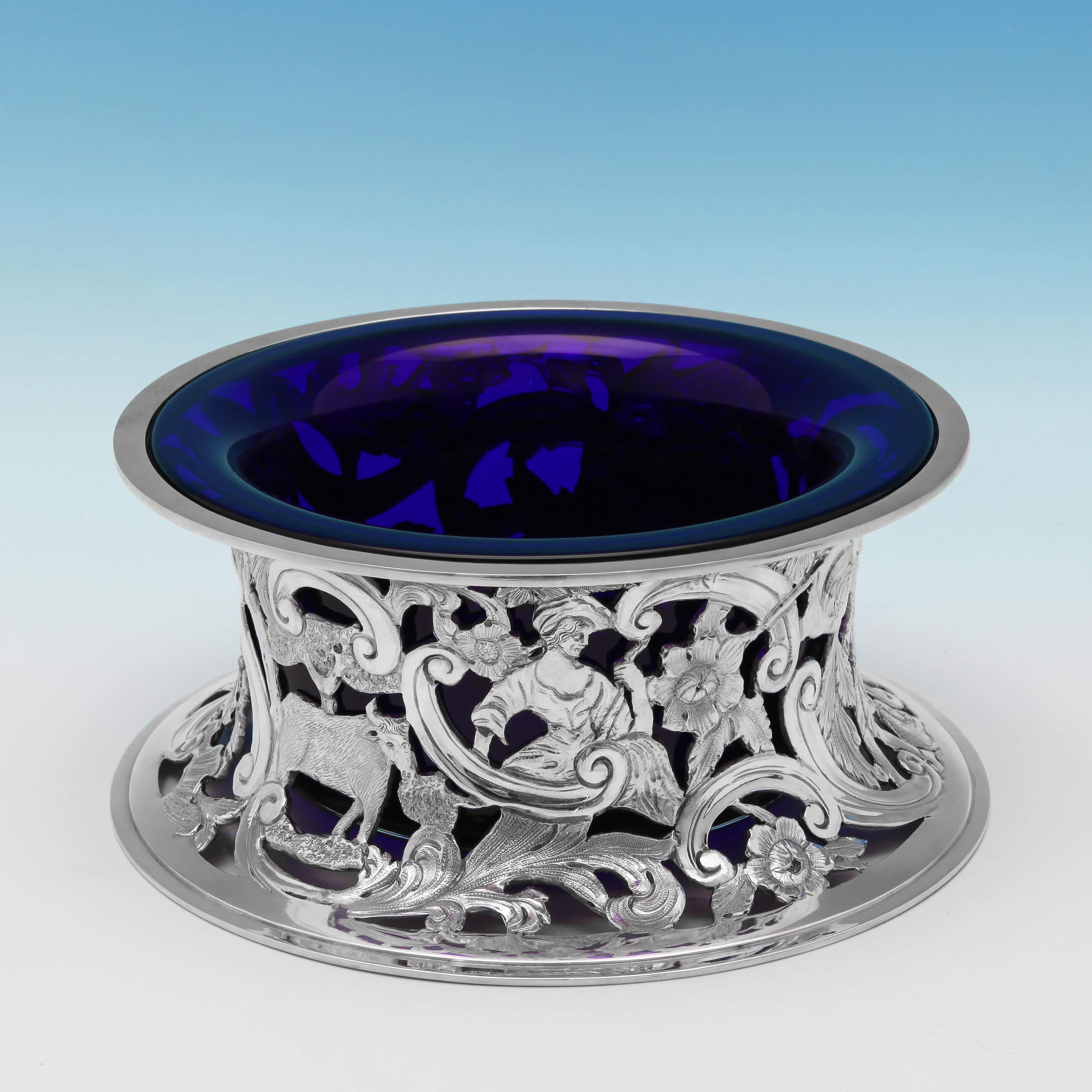 Poinçonné à Londres en 1921 par D. & J. Wellby, cet agréable anneau de plat en argent sterling présente un décor percé représentant des bergers et des animaux de la campagne ainsi qu'un décor de volutes et de fleurs, et un revêtement en verre bleu.