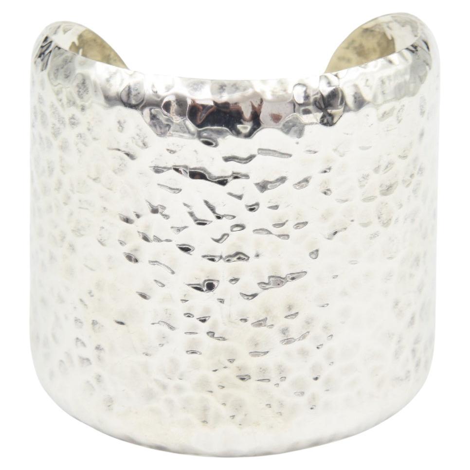 A striking cuff bracelet crafted from sterling silver.  The surface features a hammered texture, creating a dynamic play of light and shadow.  Its wide band design offers a bold statement, with a width of 2.5 inches and a height of 2.5 inches.  The