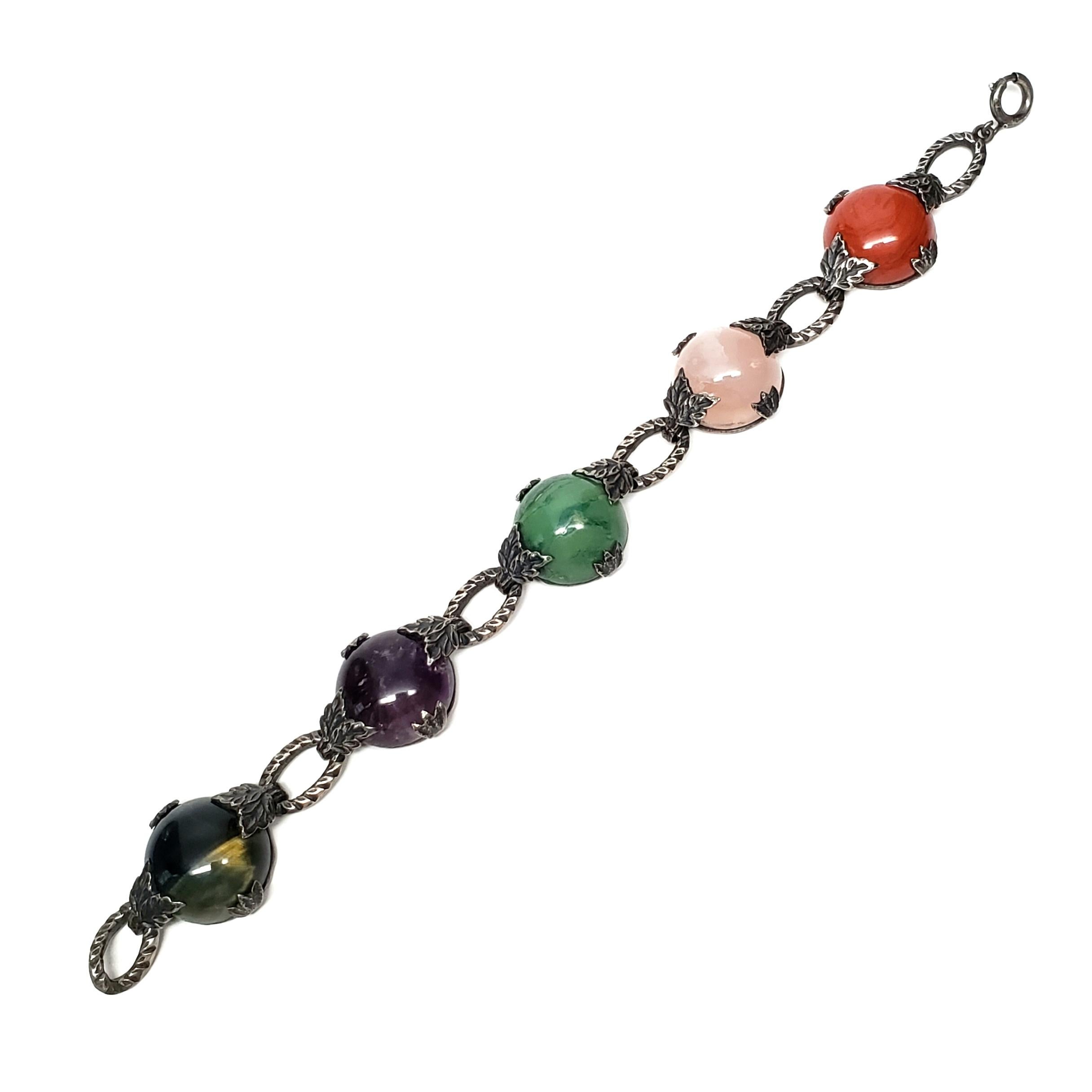 Sterling silver and multi-stone bracelet.

Features round cabochon stones set in leaf prongs, alternating with textured oval links. Stones appear to be jade, carnelian, tiger's eye, quartz and amethyst.

Measures approx 7