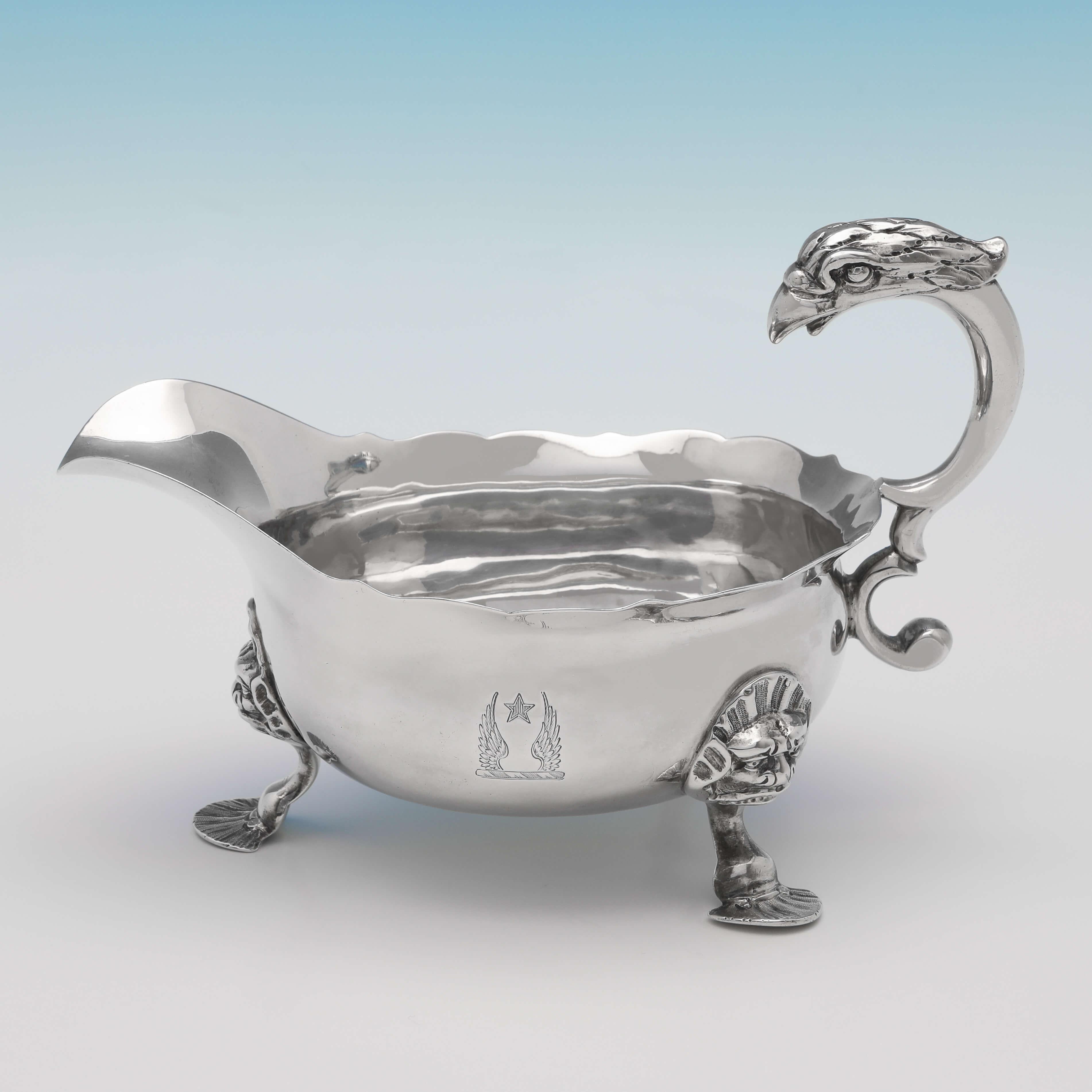 Hallmarked in London in 1748 by John Pollock, this striking pair of George II, antique sterling silver sauce boats, feature shaped borders, eagle head decorated handles, and engraved crests to both sides. Each sauce boat measures 4.75