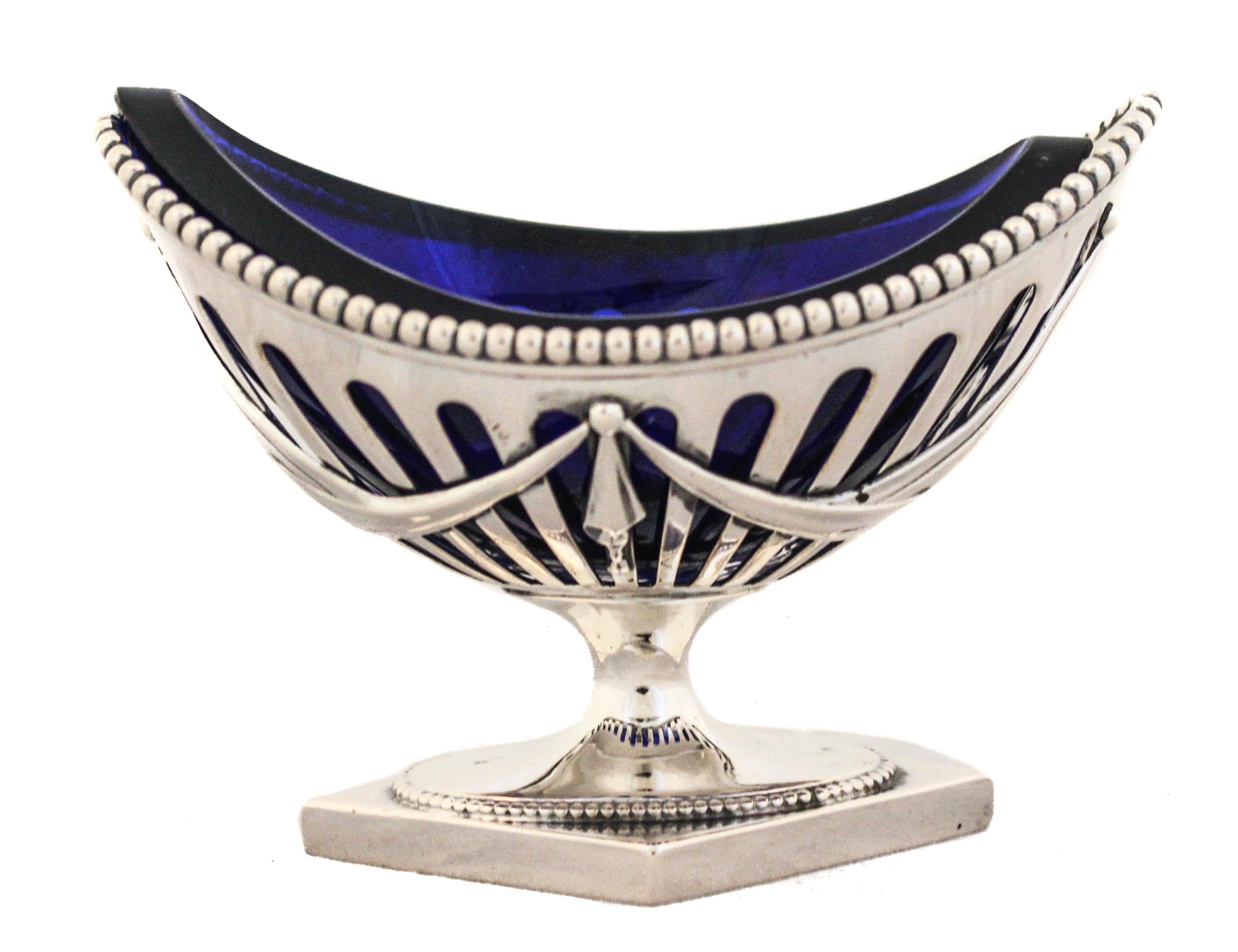 An amazing sterling silver three-piece suite: a pair of salt cellars and a matching mustard jar. Each piece has a cobalt blue liner that shows through the cutout silver. The salt cellars are oval with a diamond shaped base. A beaded design wraps