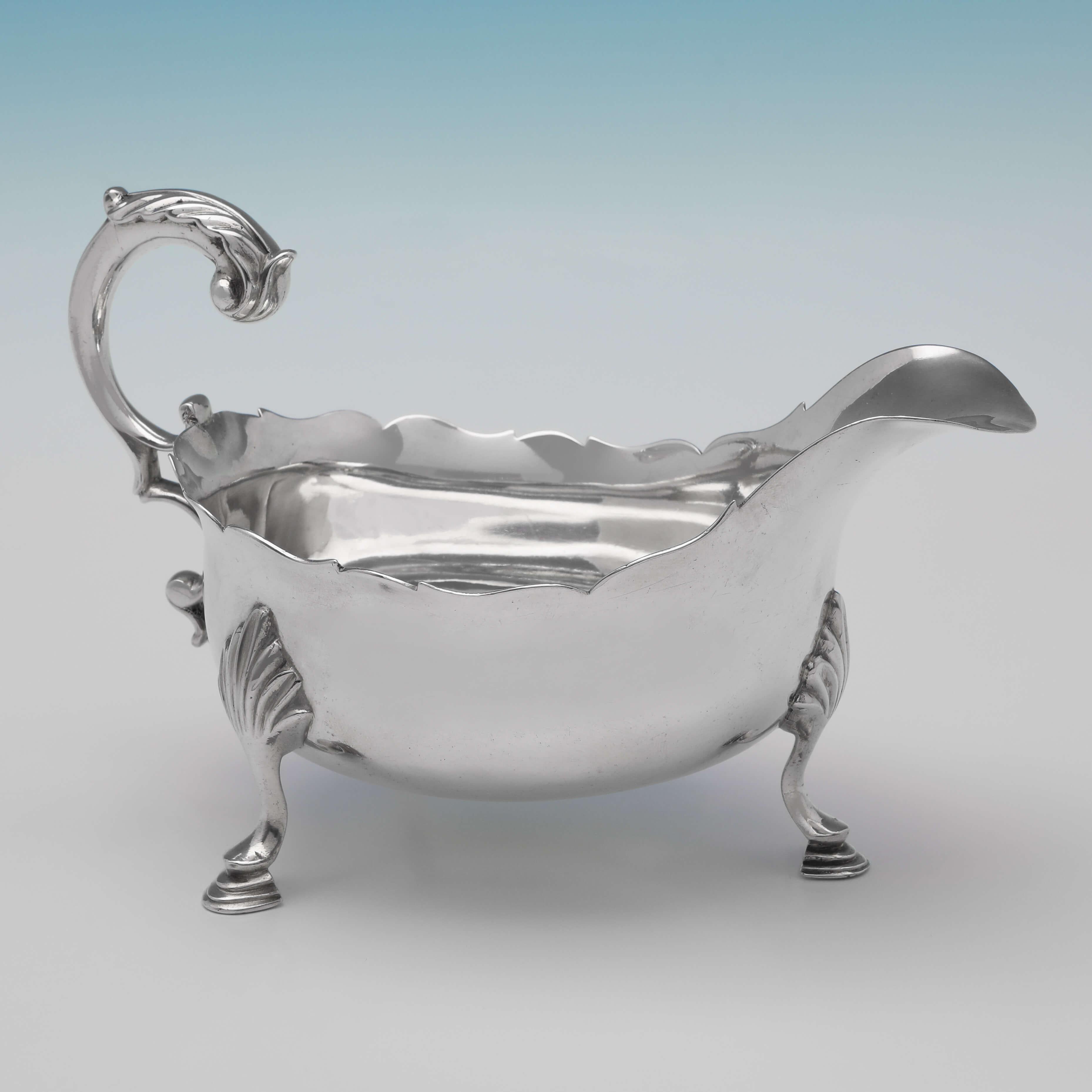 Hallmarked in London in 1760 by William Skeen, this George III, Antique Sterling Silver sauce boat, features a shaped border, an acanthus detailed flying C-scroll handle, and stands on three hoof feet. The sauce boat measures 4.5