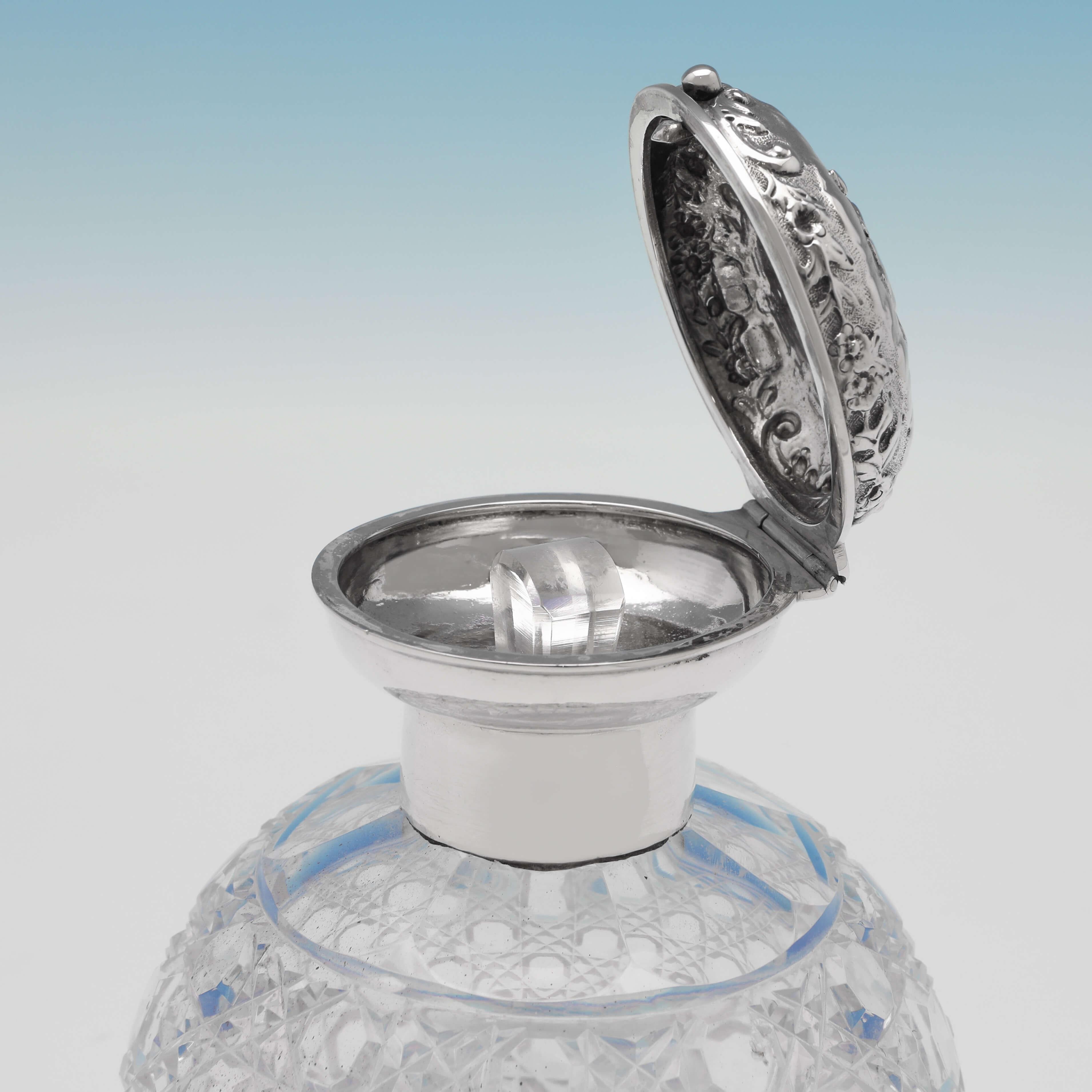 Hallmarked in London in 1902 by Mappin & Webb, this attractive, Edwardian, antique sterling silver scent bottle, features a round cut glass body, and an ornate silver lid chased with cherubs, with a push button opening. The scent bottle measures