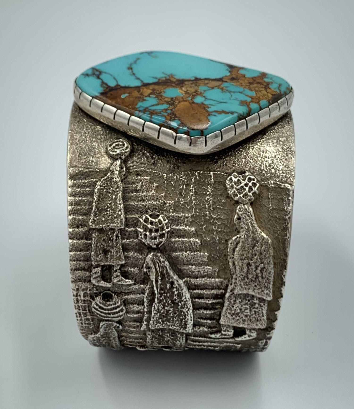Water Carrier Cuff. Sterling Silver tufa cast cuff by noted Navajo silversmiths Darryl Dean and Rebecca Begay. The 1 3/4” cuff has a beautiful 1” x 1 3/4” turquoise stone. Seven Native American water carrier women are in bas relief. Native American