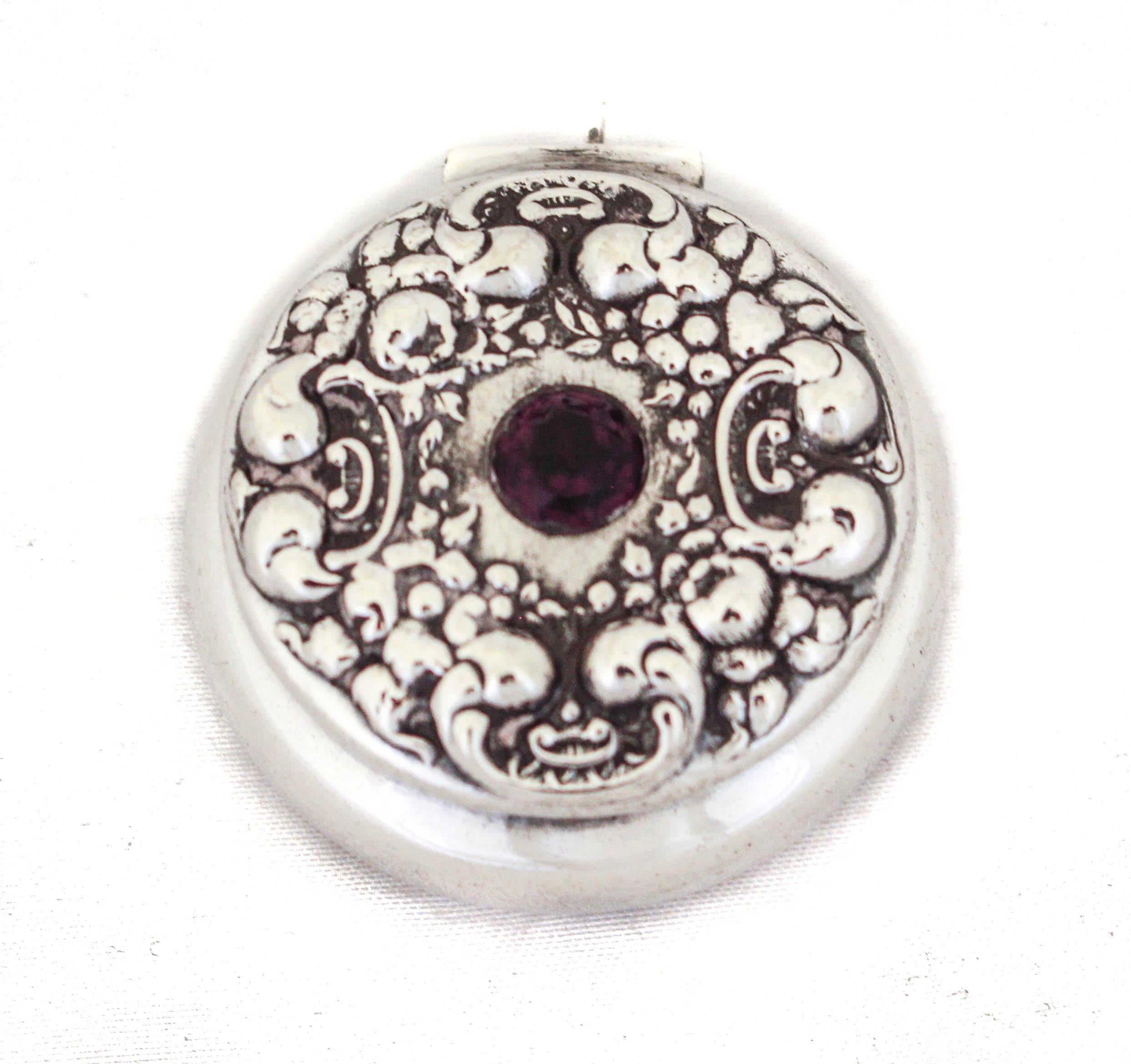Being offered is a sterling silver pillbox manufactured by the La Pierre Silver Company.   Designed at the height of the Victorian era, it has an ornate design on the top (lid) with a garnet stone in the center.  The lid is attached and opens via a