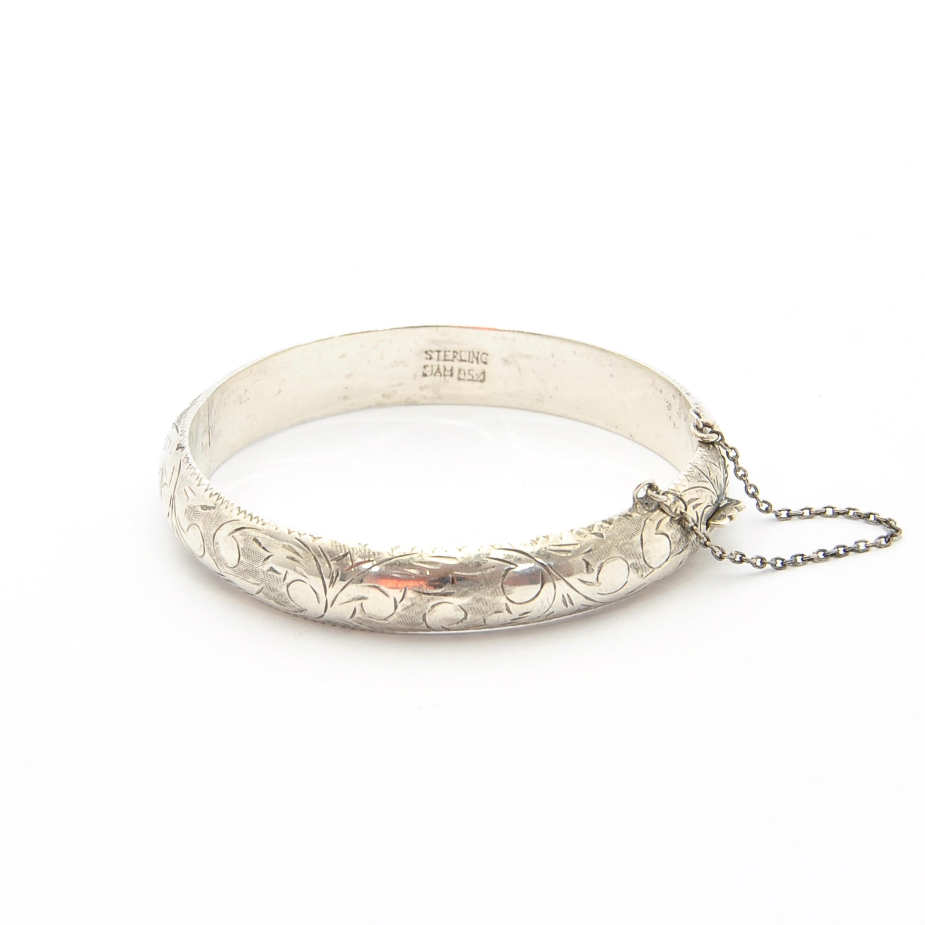 This mid-century bangle is comprised of 952 silver. It features diagonal etched lines, swirls and curl designs across its surface. The bangle has a smooth silver inside. It is hinged and closes with a push-in compression tab closure. The bracelet is