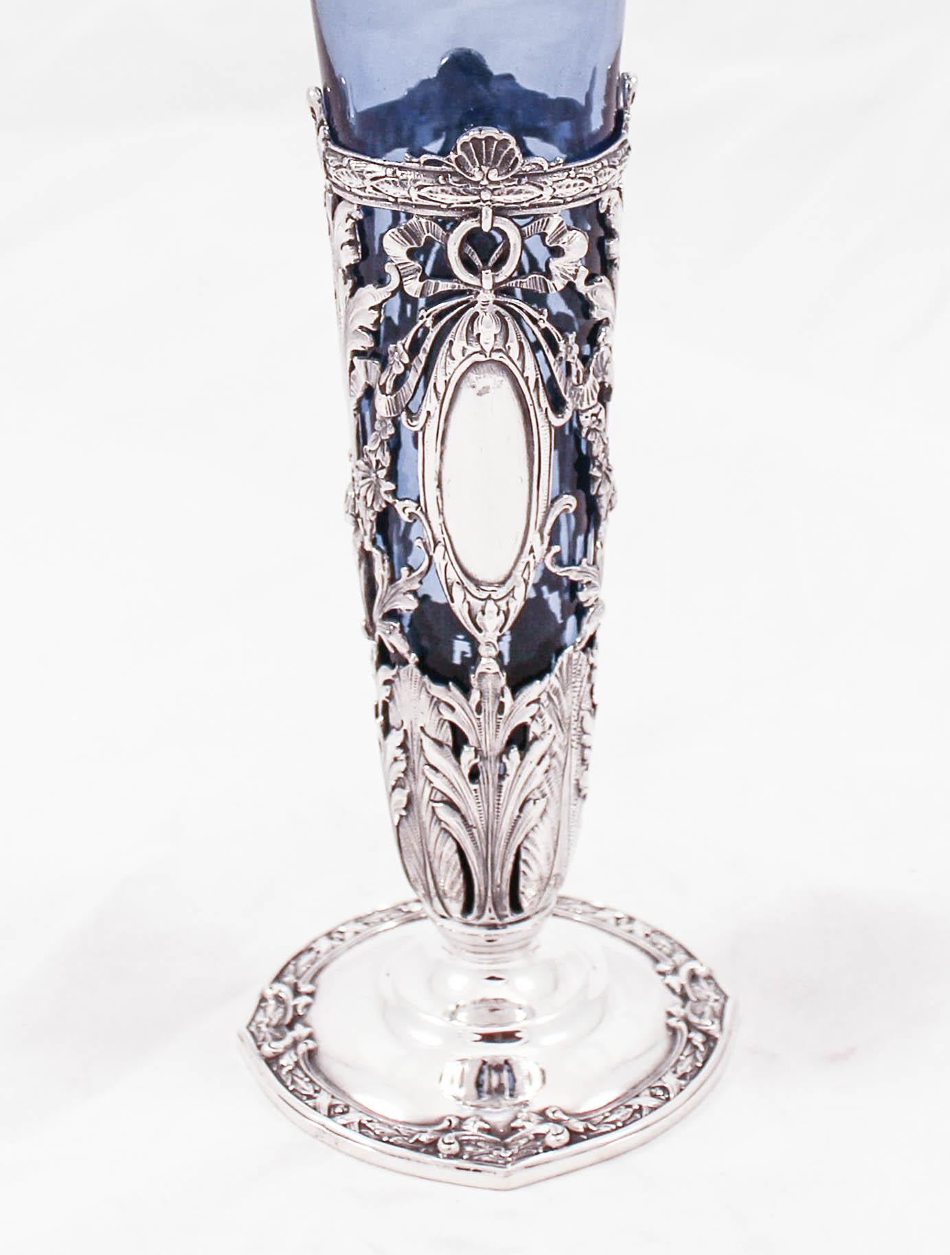 This very unique sterling silver vase has a blue glass liner. The workmanship on the sterling base is beautiful. Bows, flowers, and a cutout design gives it a rich feel and is a nod to old-world craftsmanship. The glass shows through the cutout and