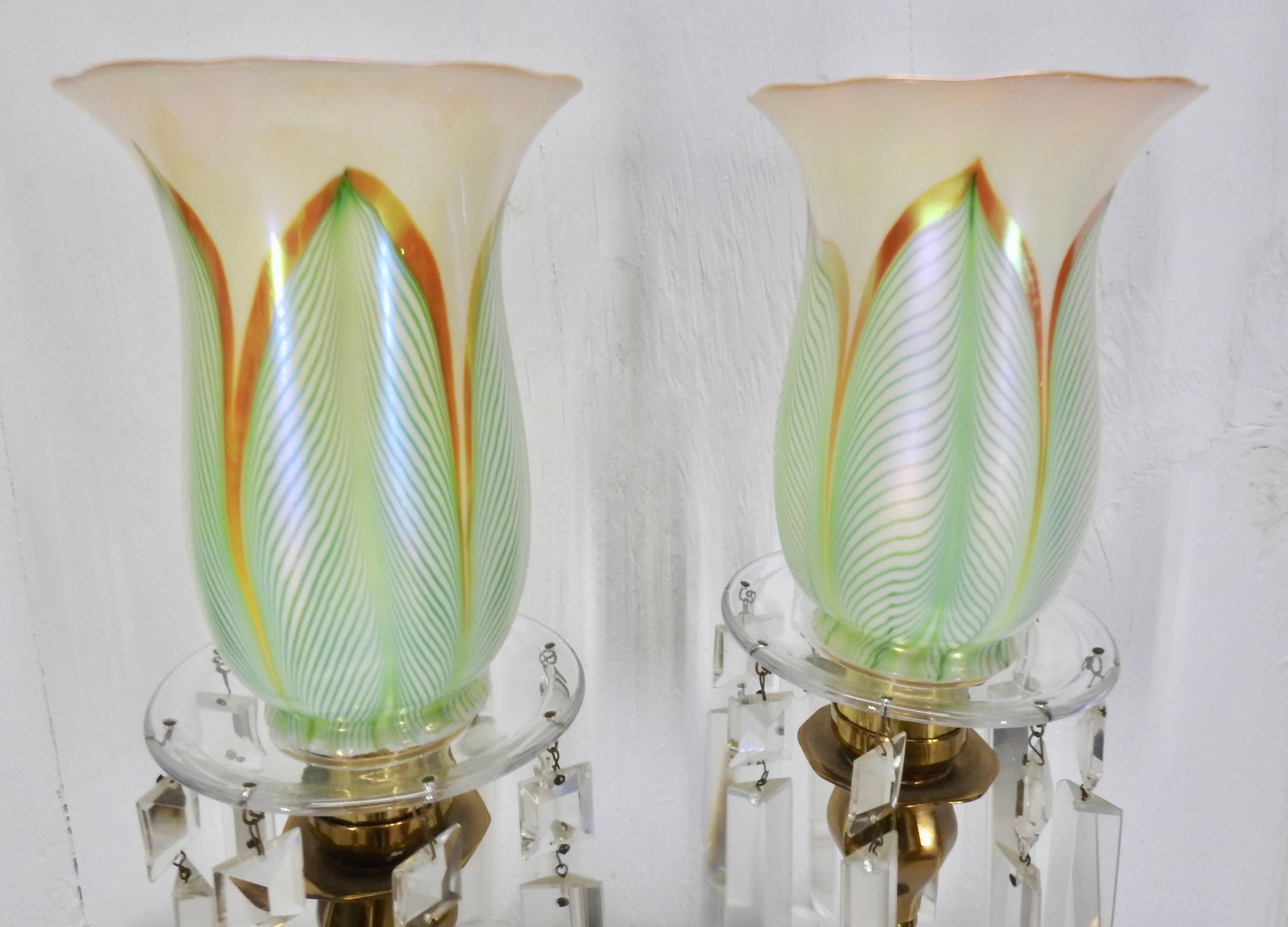 We are offering an exquisite pair of antique brass lamps with Steuben “Pulled Feather” pattern glass shades. The lamps feature a clear glass bobesche dish topping an eight-sided brass body with a raised foot. Eight faceted crystal prisms line the