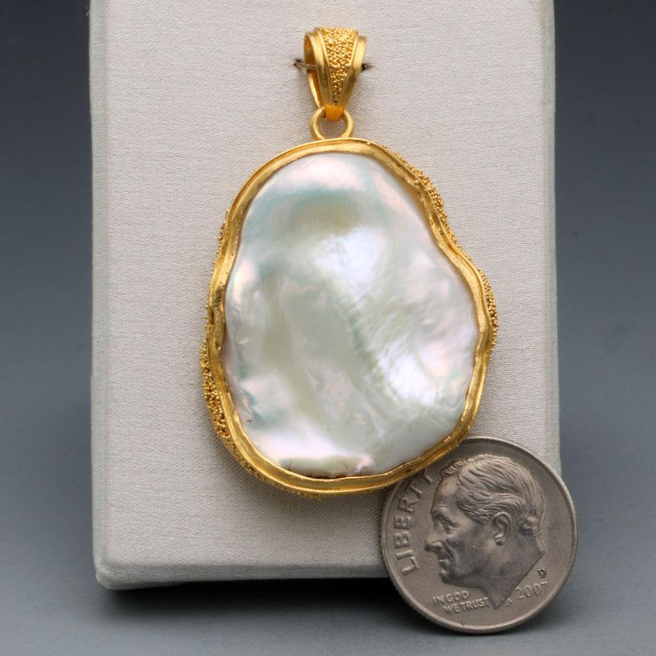 A beautiful, irregular, approximately 25 x 50 mm white large freshwater pearl with lots of lustre is set within an artisinally crafted high-karat gold bezel decorated with fine 