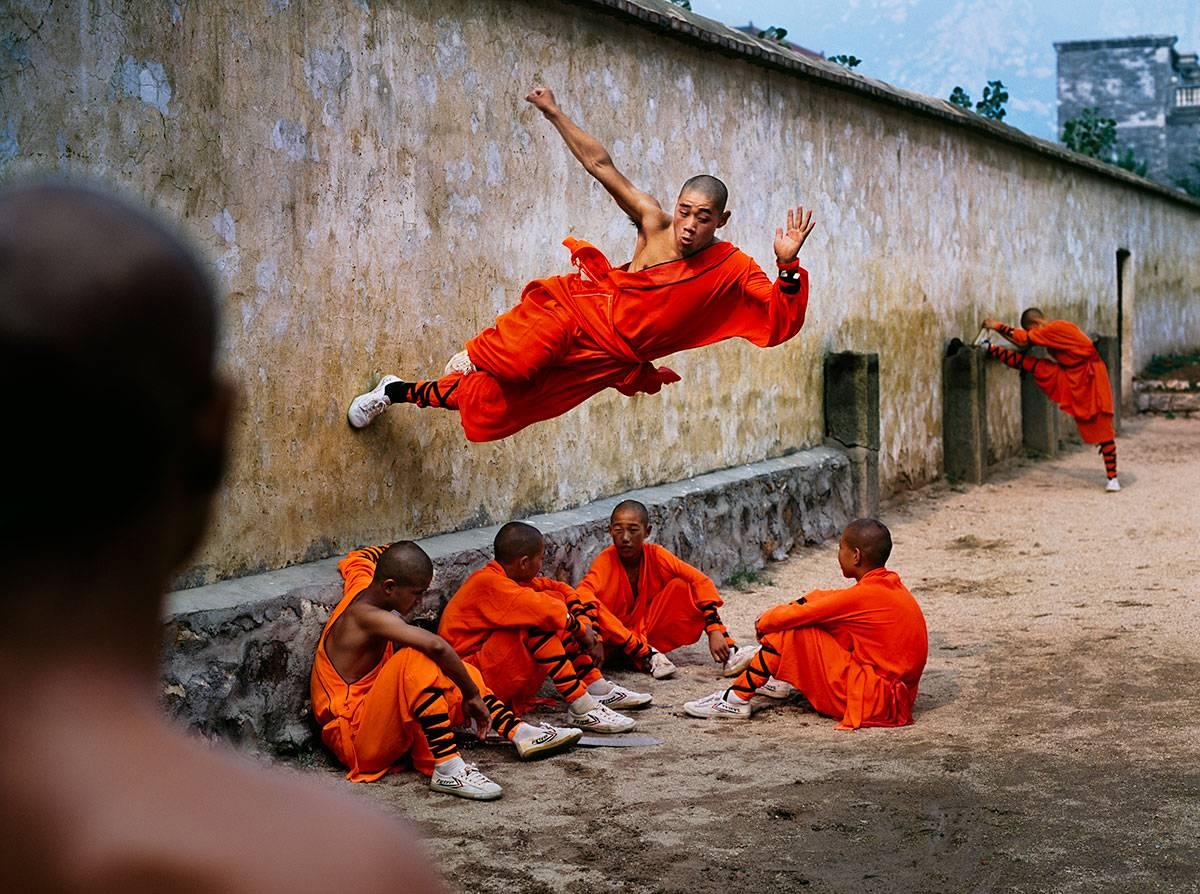 Steve McCurry Landscape Photograph - A young monk runs along the wall over his peers at the Shaolin Monastery...