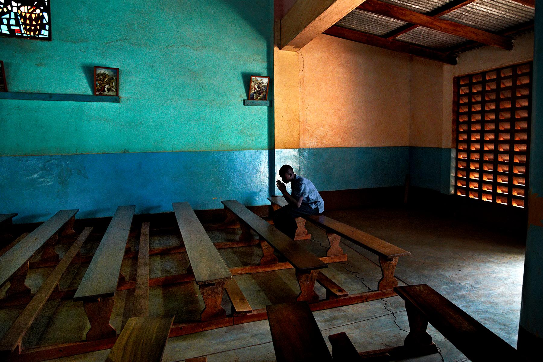 A coffee farmer prays alone in an empty church by Steve McCurry depicts a man sitting by himself on a bench, kneeling over with his hands in prayer. Light pours in from the open door, illuminating the bright blue and orange painted walls. 

This