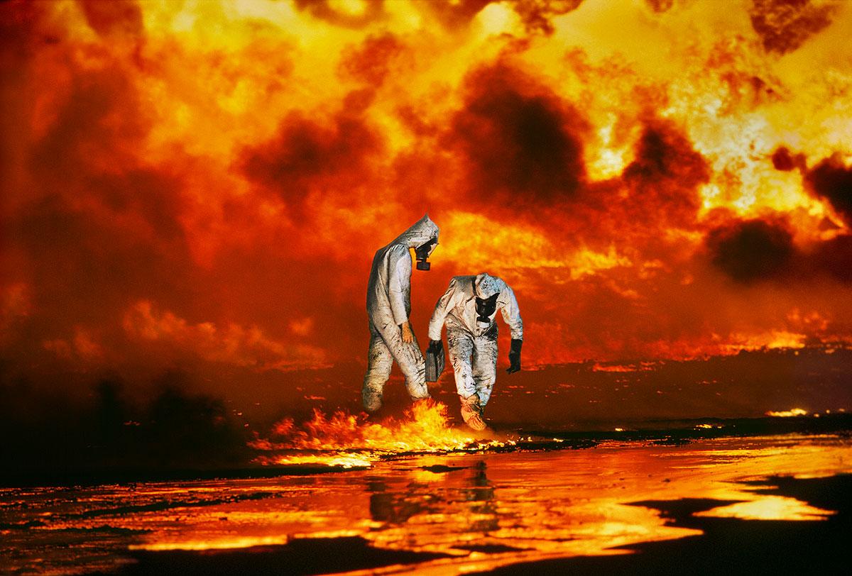 Ahmadi Oil Fields by Steve McCurry depicts two figures standing in a fiery landscape. They are both dressed in white hazmat suits with gas masks. Both figures are fixated by the fire on the ground, while the sky seems to burn behind them in vibrant