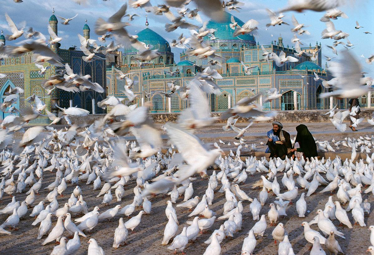 Blue Mosque, Mazar e Sharif by Steve McCurry depicts a couple seated on the ground, feeding a flock of white doves. More birds swoop down from the sky, intrigued by the hand out of food. A highly decorative blue titled mosque is visible in the