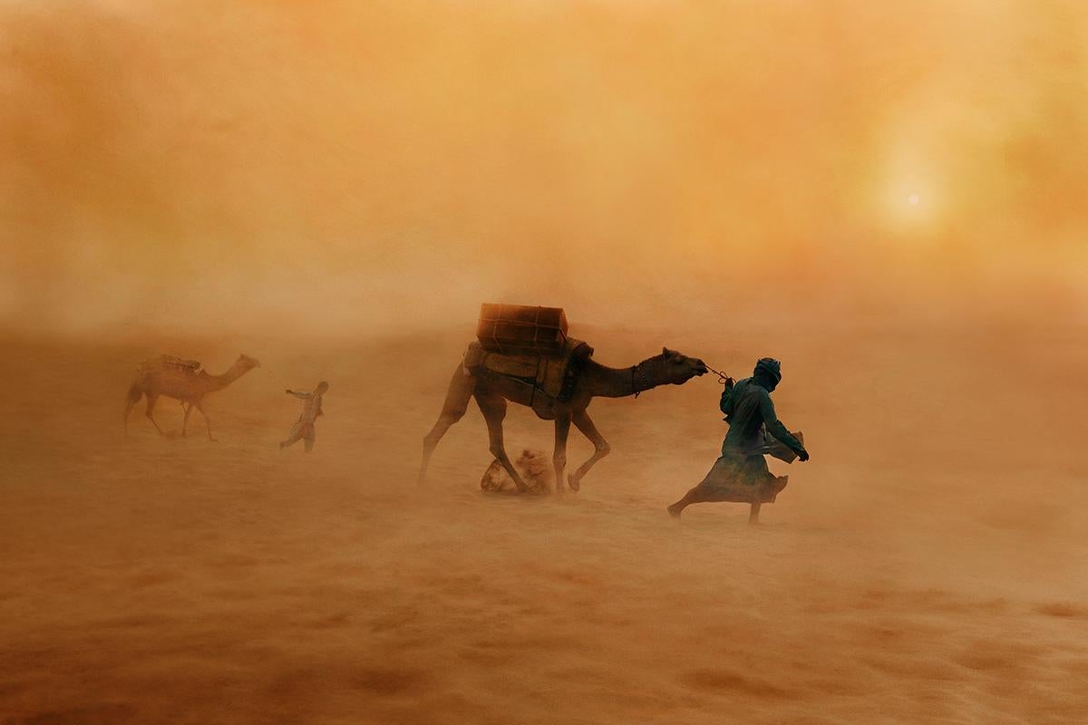Camels in Dust Storm by Steve McCurry presents two figures leading their camels through the sandy terrain of the desert. They struggle further into the storm, consumed by the orange haze of the sandy air. The sun emits a faint glow through the thick