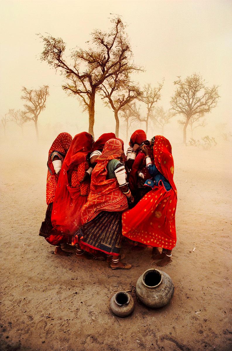 Dust Storm (Vertical) by Steve McCurry is a 30 x 24 inch Digital C-Print on FujiFlex Crystal Archive Supergloss Paper, available in an edition of 75. This photograph features a group of women, all wearing red, huddled together amidst a dusty, barren