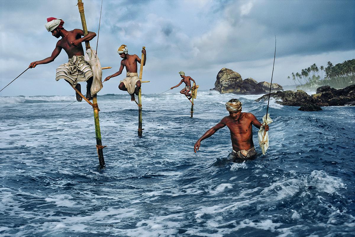 Fishermen at Wligama by Steve McCurry features four men fishing in the ocean, on the shores of Sri Lanka. Three of the men are sitting on top of tall sticks, perched above the water while they cast their fishing lines below. Another man wades