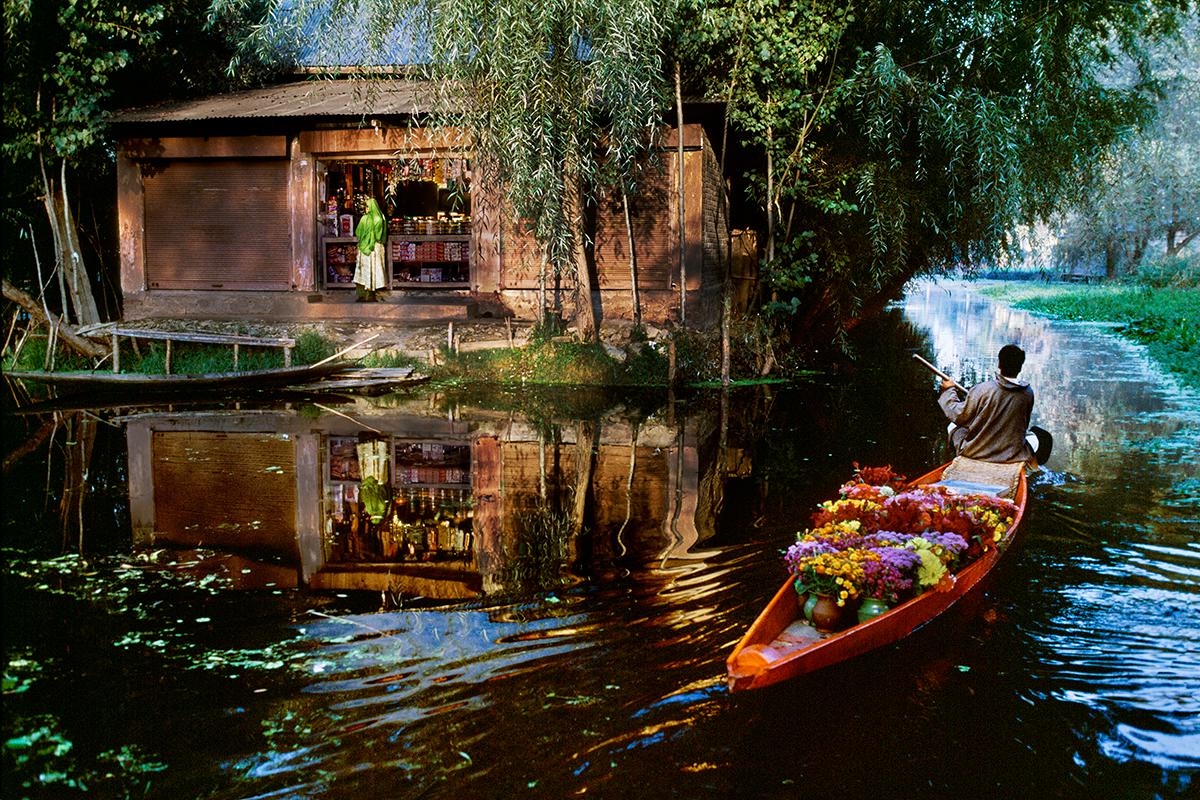 Flower Vendor on Dal Lake von Steve McCurry zeigt einen Mann, der farbenprächtige Blumen in einem Kanu transportiert. Er paddelte den ruhigen Fluss hinunter, vorbei an einem Haus, das von üppiger Vegetation umgeben war. Eine grün gekleidete Frau