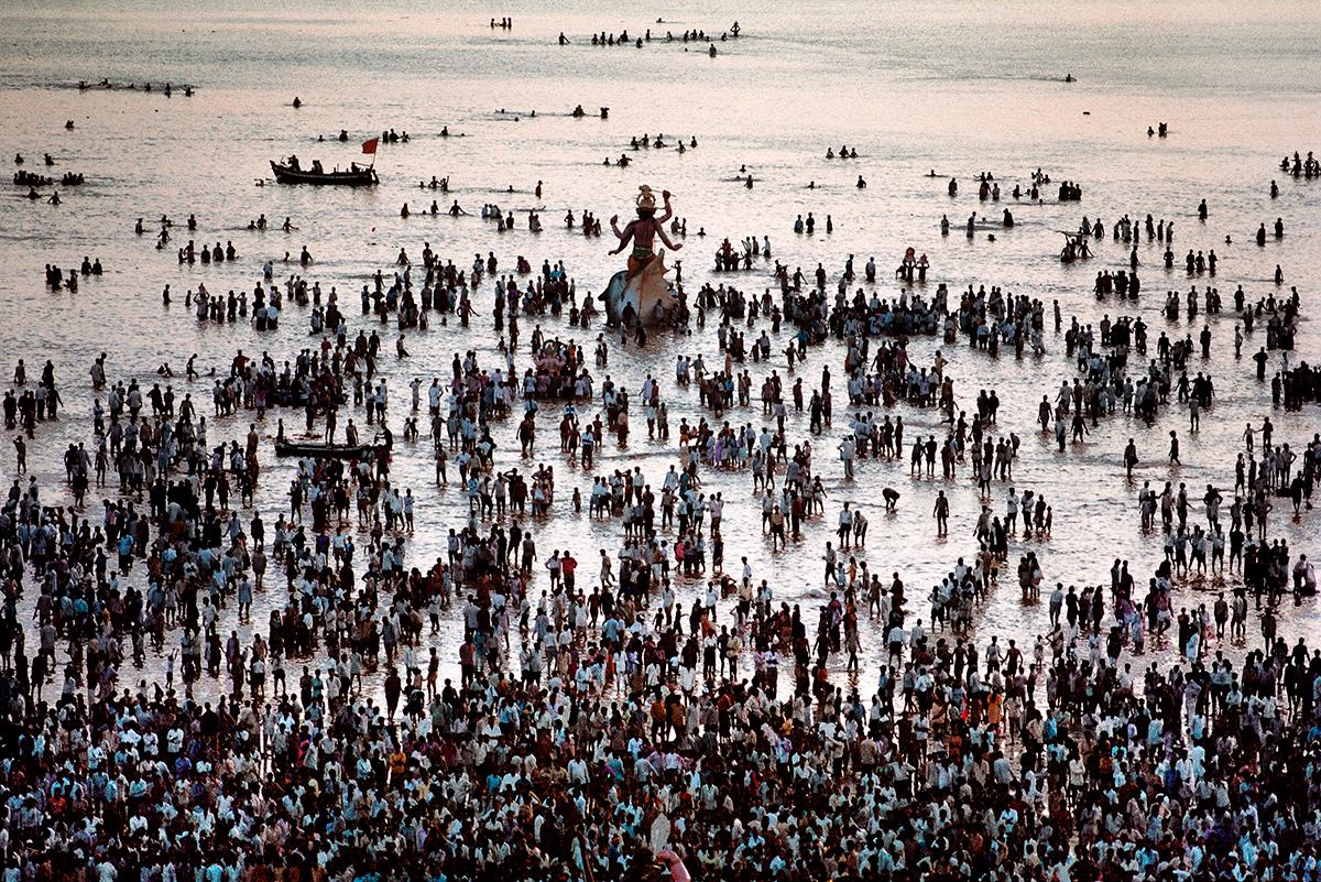 Ganesh Chaturthi Festival by Steve McCurry features a mass of people walking into the shallow waters of the sea, as part of their celebration. People gather around a large statue of the Hindu god Ganesha as they continue to walk towards the