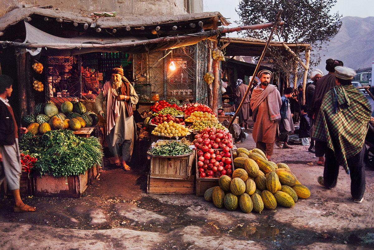 Harvest Market by Steve McCurry is a 20 x 24 inch digital C-print, printed on FujiFlex Crystal Archive Supergloss Paper. This photograph features a man standing in a market, surrounded by various colorful fruits. This photograph is signed by Steve