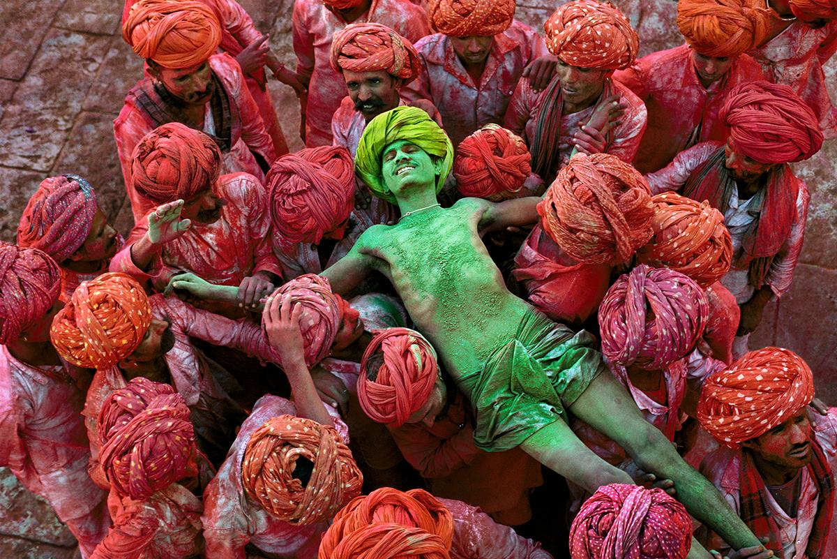 Holi Man von Steve McCurry zeigt eine Gruppe von Männern während des hinduistischen Holi-Festes. Ein mit grünem Pulver bedeckter Mann liegt auf dem Rücken, während ein Meer von roten Männern ihn hochhebt und trägt. Auf den Gesichtern der Männer sind