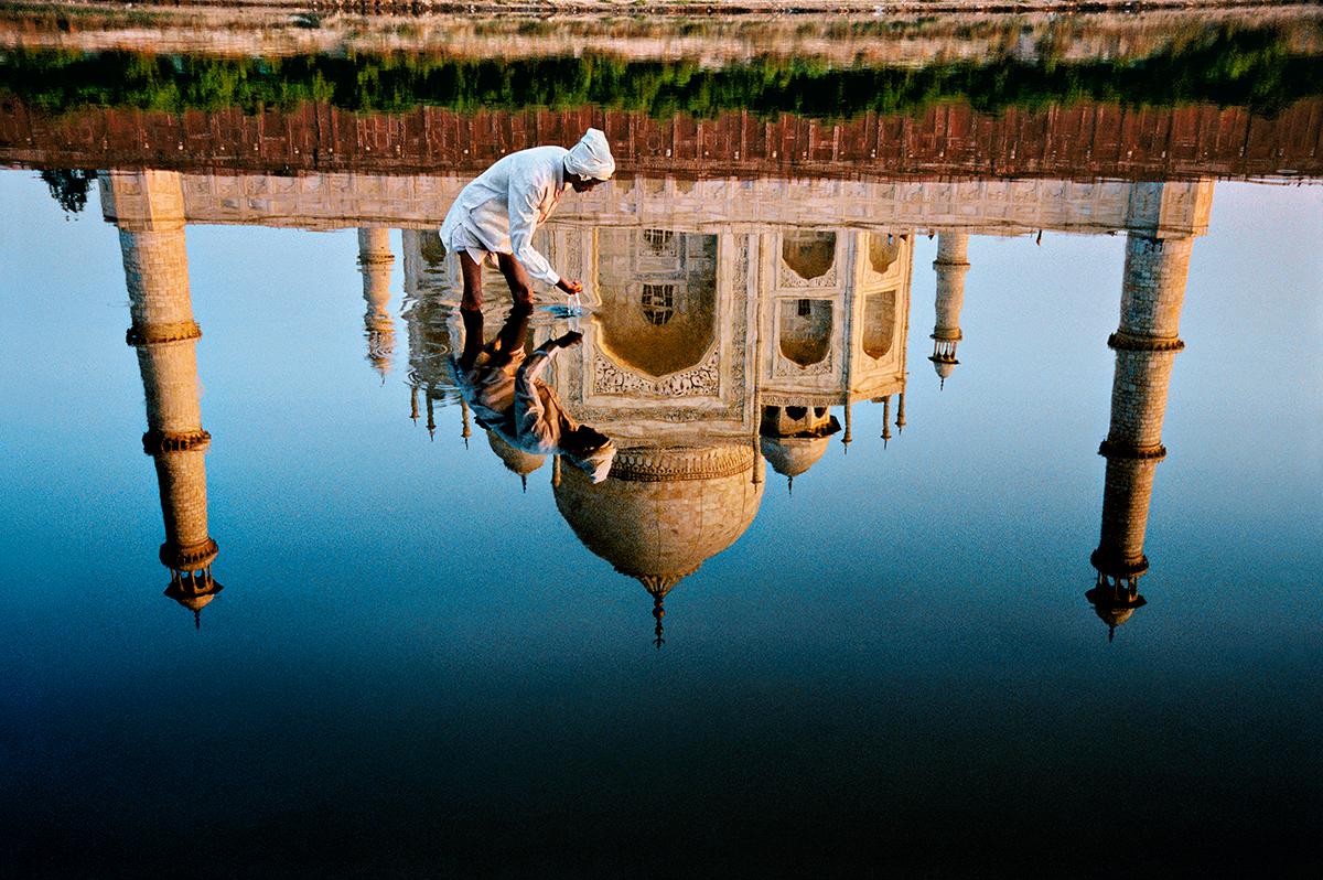 Man and Taj Reflection von Steve McCurry zeigt einen weiß gekleideten Mann, der sich hinkniet, um Wasser unter sich zu sammeln. Das Taj Mahal spiegelt sich fast perfekt im stillen Wasser.

Diese Fotografie ist ein digitaler C-Print, gedruckt auf