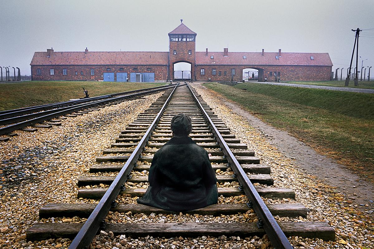 Man Meditates at Auschwitz von Steve McCurry zeigt einen Mann, der in der Gedenkstätte und Museum Auschwits-Birkenau in Polen mitten auf den Gleisen sitzt. Die Gleise führen zurück zu den Toren des großen roten Backsteingebäudes im Hintergrund. Das