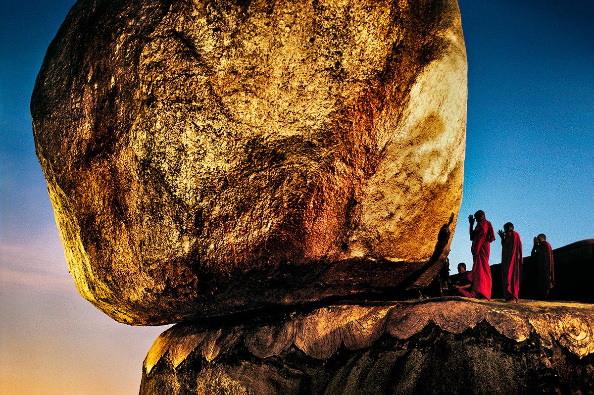 Monks Praying at Golden Rock by Steve McCurry presents a group of men praying next to a giant rock. The four men are dressed in red, with their hands in prayer raised to their forehead. They are dwarfed by a colossal boulder, bathed in golden light,